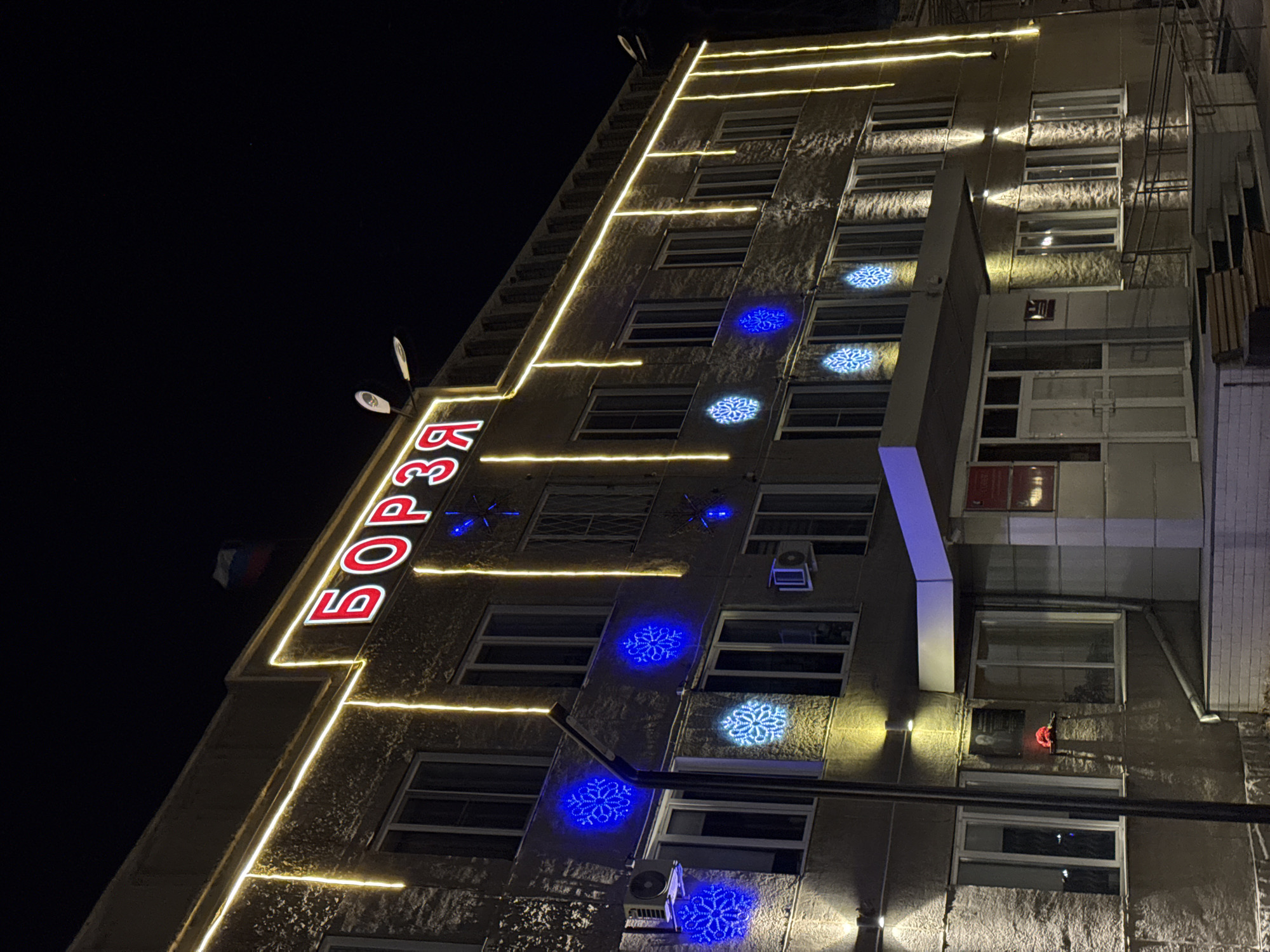 The image depicts a tall building in Borzya, Russia, illuminated at night. The name "БОРЗЯ" is vertically displayed in red neon lights along the building's left edge. Horizontal yellow string lights accentuate each floor. Blue snowflake light projections adorn the building's facade between the windows. The building has multiple floors with windows visible. A satellite dish and a Russian flag are visible on the left side of the building.