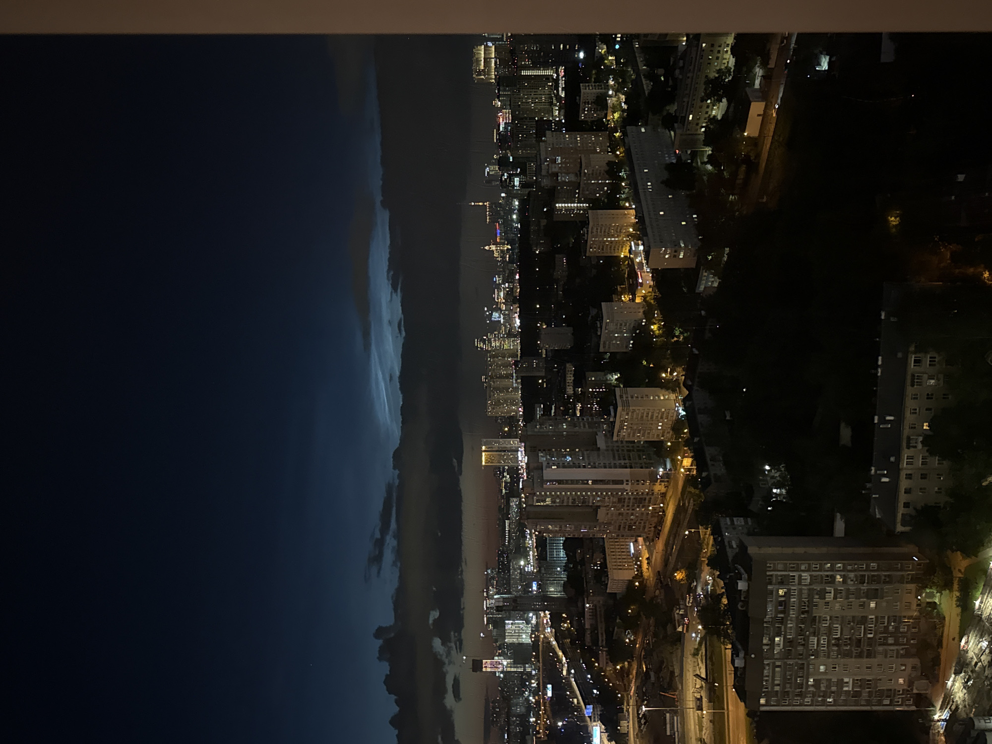 The image depicts a nighttime aerial view of a city. The scene is divided into two distinct halves by a dramatic weather phenomenon. On the left, a dark, stormy sky looms with visible clouds. The right half of the image reveals a brightly lit cityscape, characterized by numerous buildings illuminated by artificial lights. The architecture suggests a dense urban environment with residential and possibly commercial structures. Roads and smaller illuminated objects, likely vehicles, are also visible, indicating ongoing activity. The time of day is clearly night, with the darkness of the sky contrasting sharply with the man-made illumination of the city. The weather appears to be transitioning from stormy to clear, or vice versa, as suggested by the cloud line. There is no visible text in the image. The overall impression is one of a dynamic urban environment under a dramatic sky.