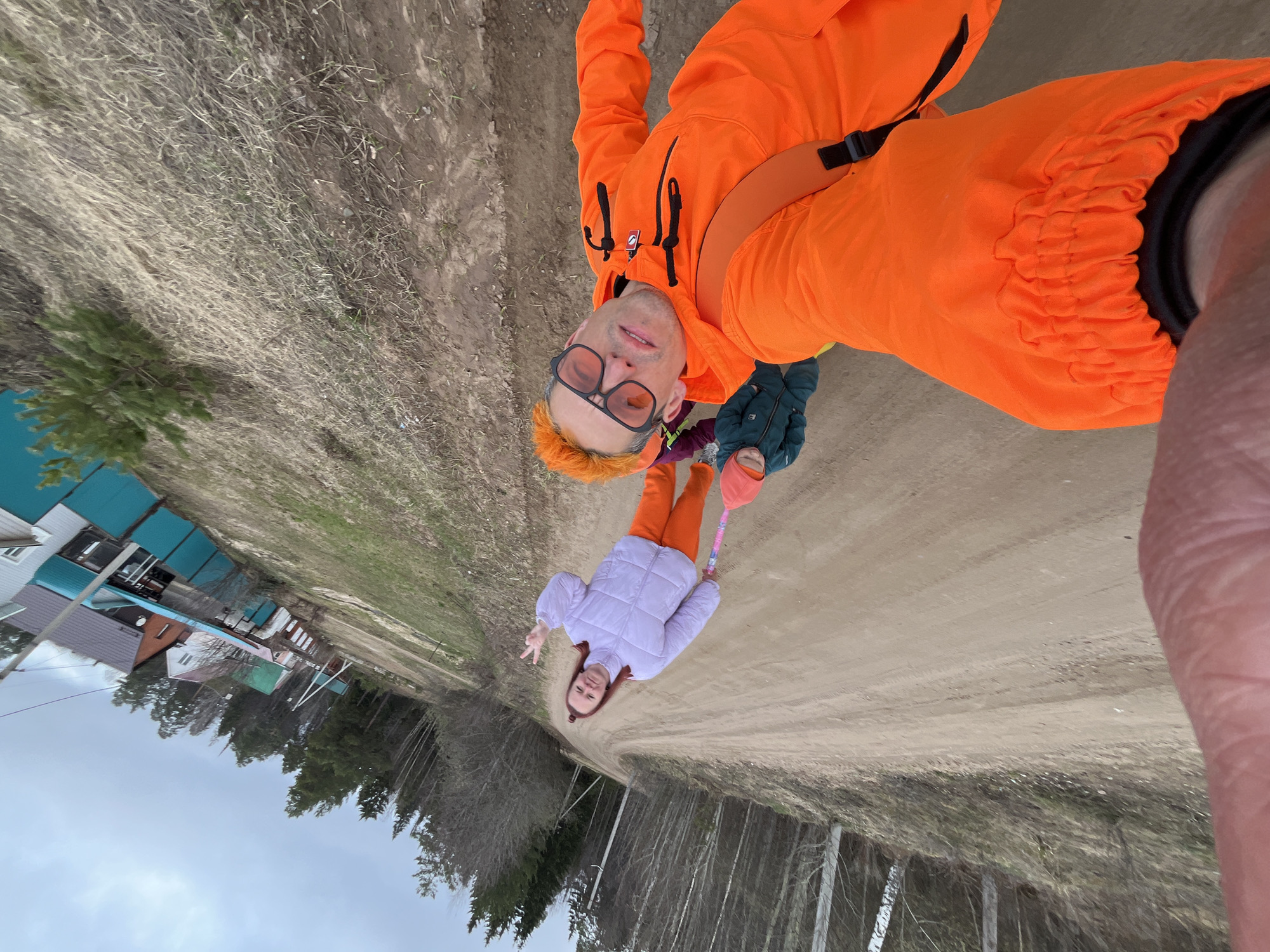 An outdoor, rotated image, set in Chaikovsky, Russia, features three individuals on a dirt road, likely during a casual outing or walk. The main subject, an adult with orange-dyed hair and orange-tinted glasses, is wearing a bright orange jacket and matching pants. This person is holding the camera for what appears to be a selfie, looking directly at the viewer with a slight smile. A strap, possibly from a camera or bag, is visible across their chest.
To their left, a child in a light purple puffer jacket and orange pants makes a peace sign towards the camera. Part of another small child or baby, dressed in blue-green clothing, is visible between the adult and the first child, seemingly being held or standing very close.
The setting is a rural or semi-rural unpaved road, with dry, sparse grass and bare deciduous trees lining the sides, suggesting early spring. In the background, several houses or cottages are visible, some featuring teal-colored roofs or coverings. The sky is overcast, indicating a cloudy day, possibly cool given the jackets worn by all subjects. No visible text is present in the image. The overall mood appears casual and relaxed.