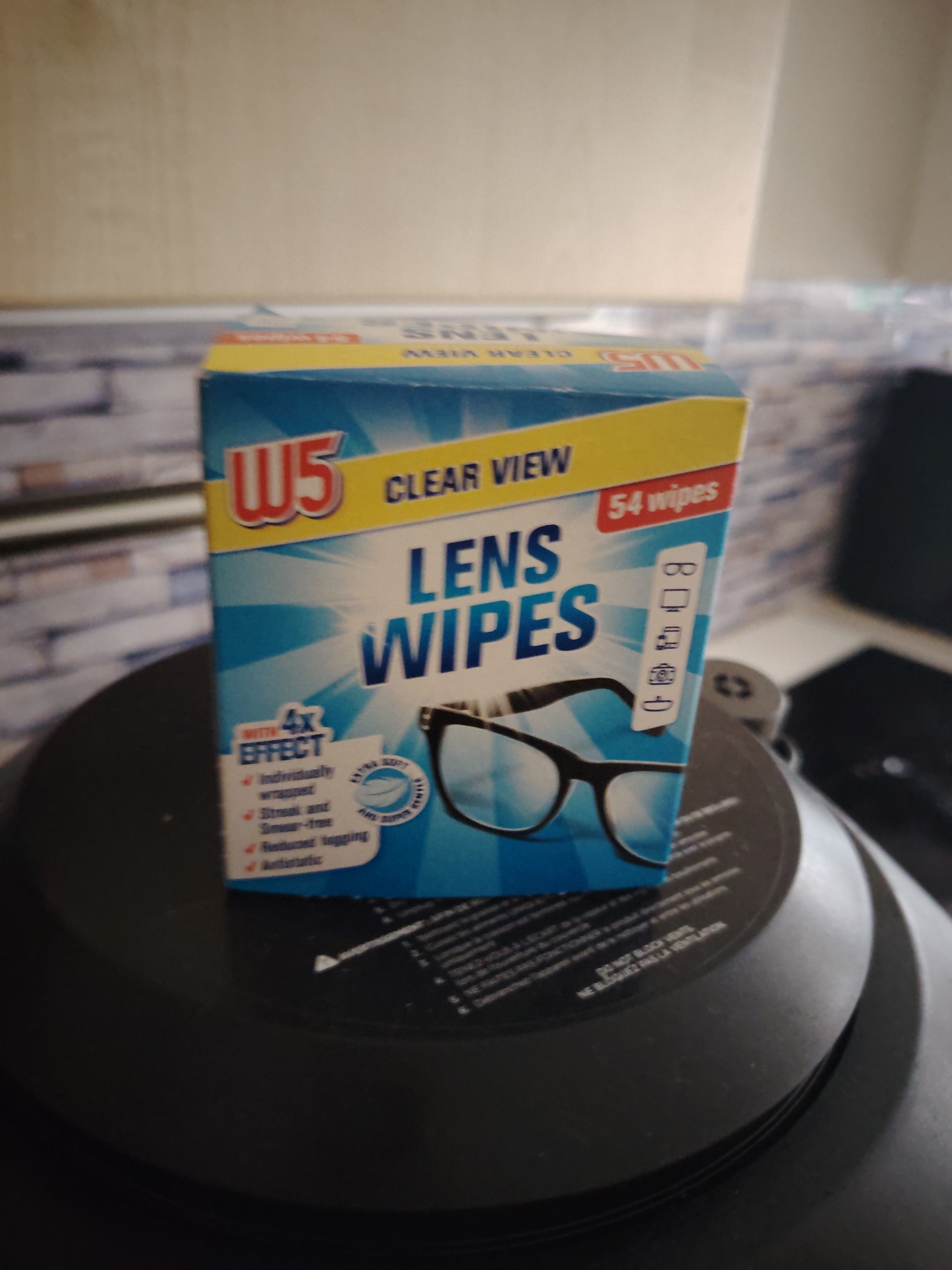 A box of "W5 CLEAR VIEW LENS WIPES" is positioned upright in the foreground of the image. The box features a blue and white design, a yellow top banner, and red accents for the "W5" logo and "54 wipes" count. The front panel displays a graphic of black-framed eyeglasses and lists features such as "WITH 4X EFFECT," "Individually wrapped," "Streak and Smear-free," "Reduces fogging," and "Antistatic." Icons illustrating suitable uses for the wipes include glasses, a computer monitor, a tablet or smartphone, a camera, and binoculars. The box rests on a dark, multi-tiered circular object, possibly the base of a small appliance, with faint white text visible on its surface, including phrases like "DO NOT BLOCK VENTILATION." In the background, to the left, a light-colored wall with a subtle textured pattern, resembling brickwork, is visible. Behind this and extending across the top, a plain, light-colored wall or surface appears. To the far right, a dark electronic device, possibly a speaker or part of a computer, is partially visible. There are no people present, and no actions or interactions are depicted. The specific location within Huntingdonshire, United Kingdom, cannot be determined from the visual information provided in the image.