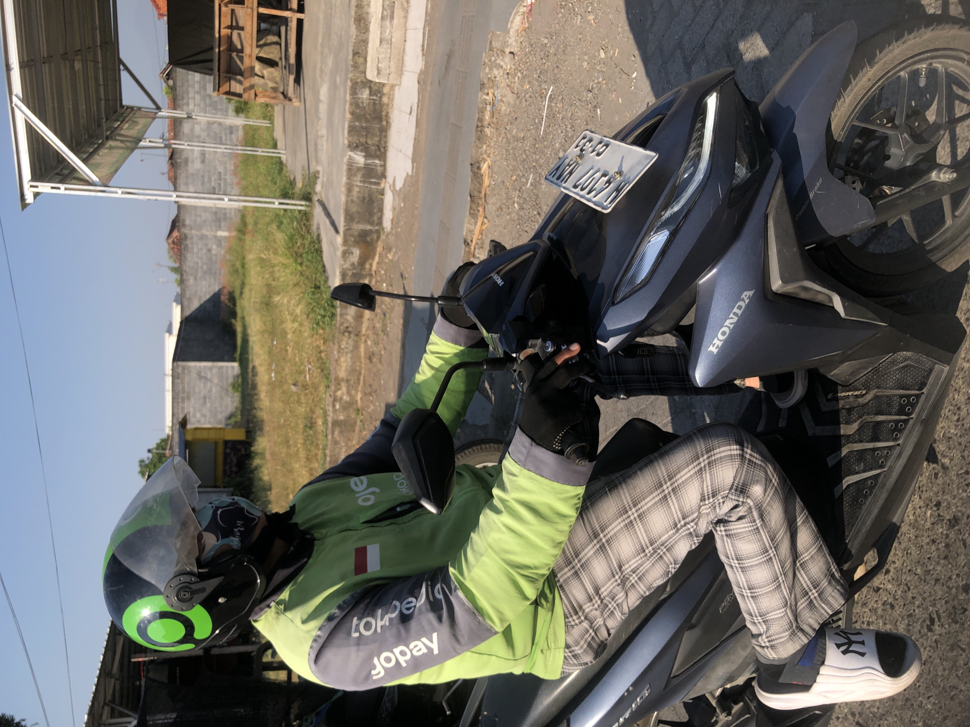 A person is riding a dark grey Honda motorcycle. The rider is wearing a black helmet with a green logo, a light green and grey jacket with "Gojek" branding, black gloves, and plaid pants. The motorcycle has a license plate that reads "N 4107 WAX CJ-53". The scene appears to be outdoors on a sunny day with a clear blue sky. In the background, there is a concrete wall, some dry grass, and a small building structure with a metal roof. The location is identified as Boro, Indonesia.