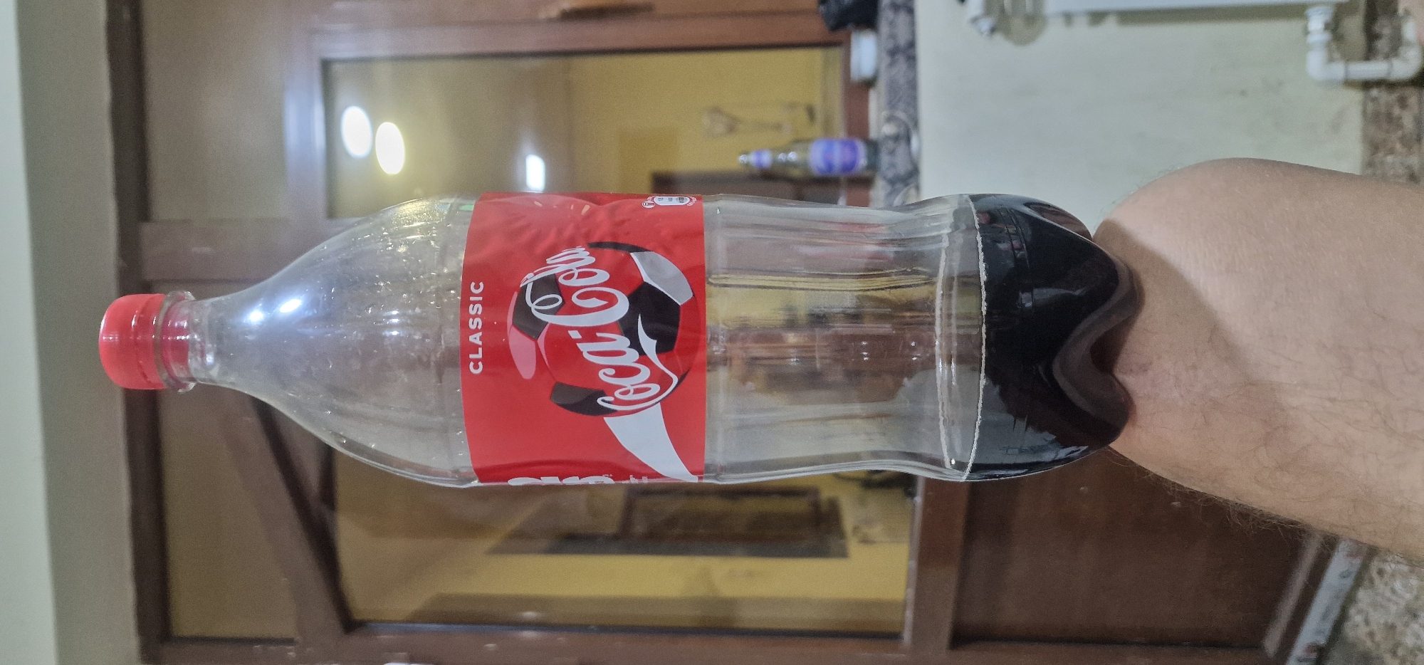 A clear plastic Coca-Cola bottle, approximately half full of dark liquid, is balanced on top of a person's leg. The bottle has a red cap and a red label featuring the classic Coca-Cola logo with a soccer ball graphic and "CLASSIC" text above it. The background is a room with wooden doors and windows, suggesting an indoor setting. The lighting is dim, with some reflections visible on the bottle and the background. There is no specific indication of the location being Karshi City, Uzbekistan.