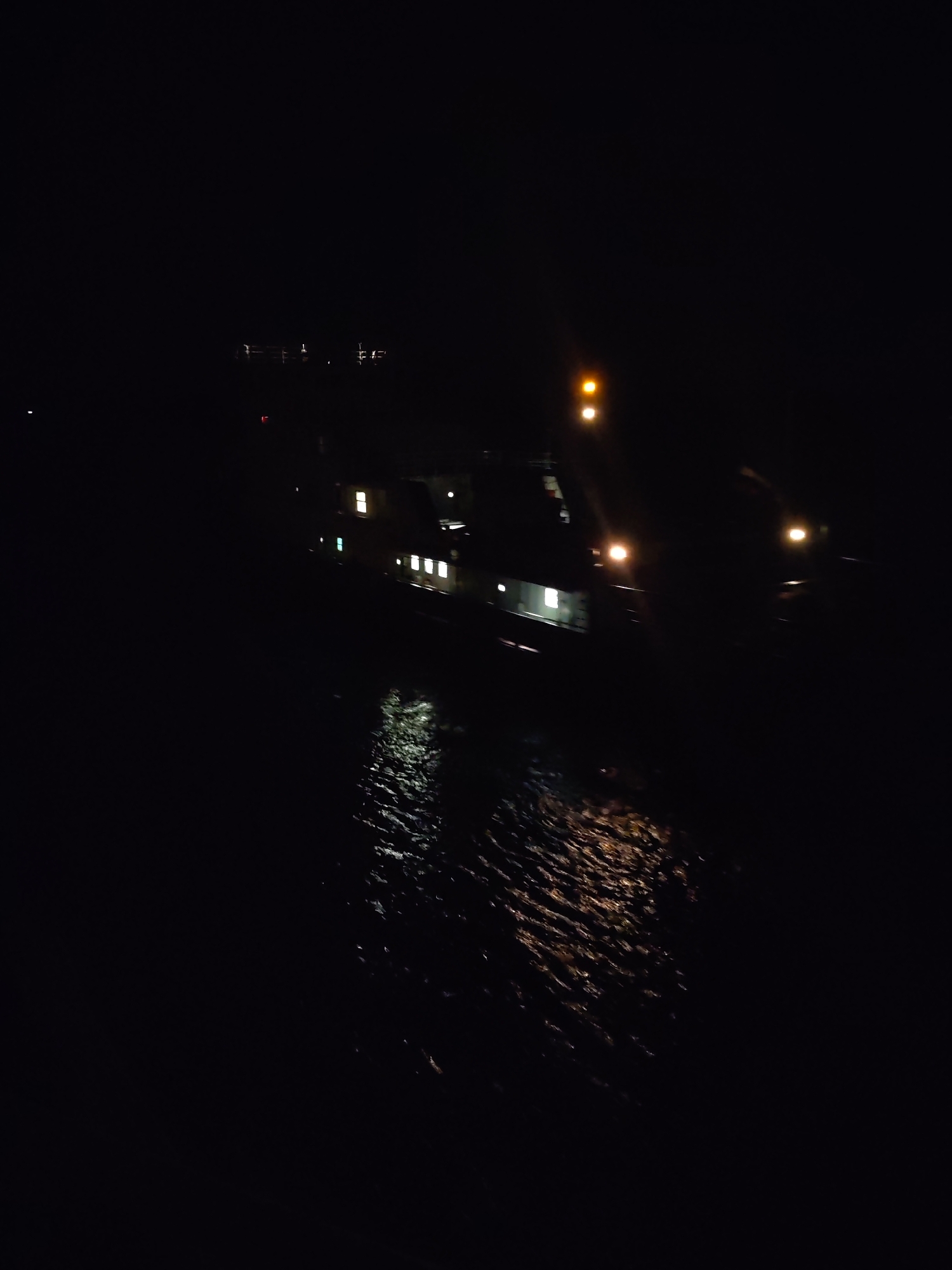 The image depicts a night scene in Gorka, Russia. A body of water reflects light from a vessel with multiple illuminated windows and running lights. A single, brighter light source is visible higher on the vessel. In the distance, faint lights suggest a city skyline. The foreground is predominantly dark, indicating the viewer is positioned at an elevated vantage point overlooking the water. The water's surface shows ripples and reflections, suggesting slight movement.
