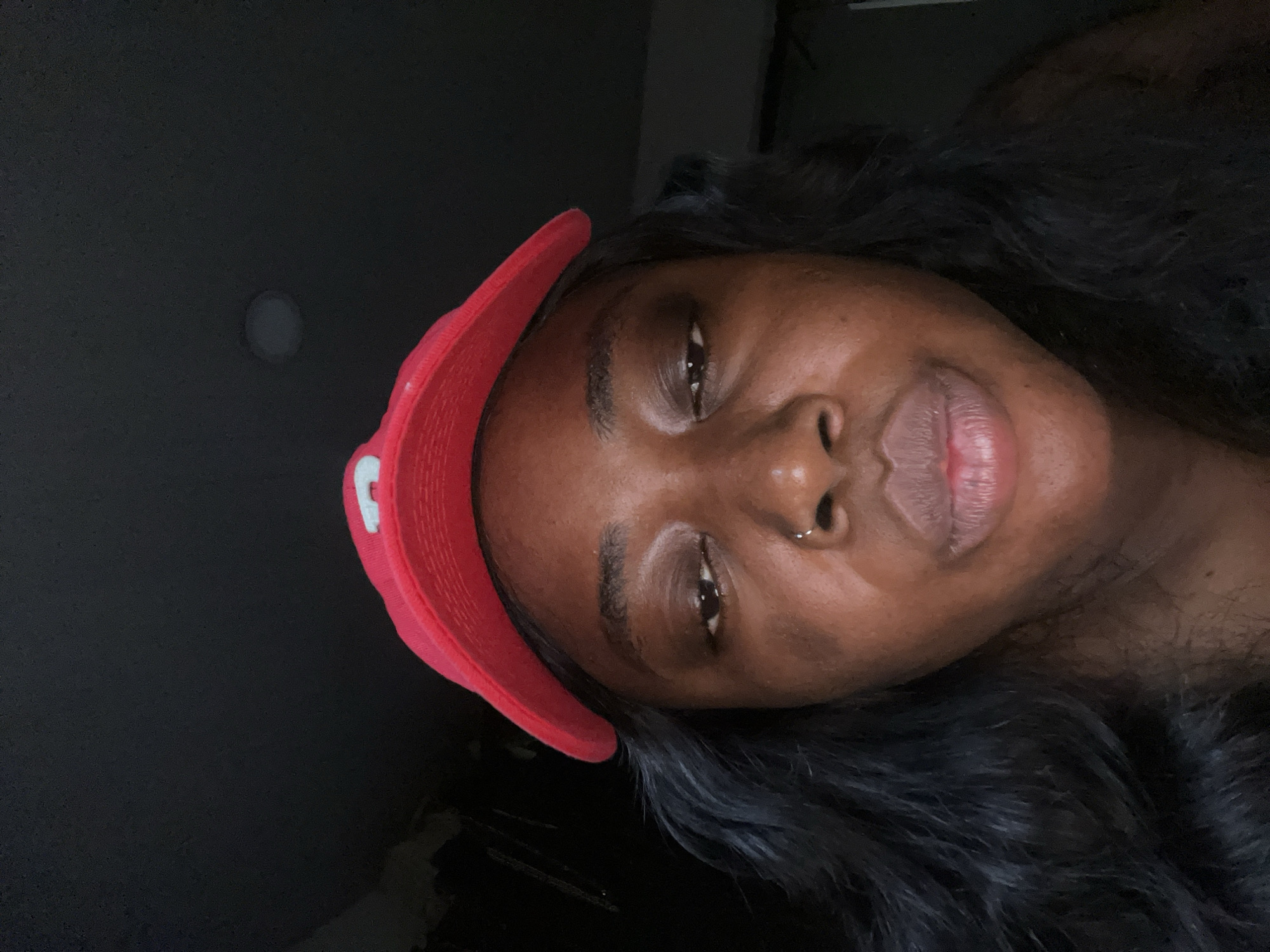 The image is a close-up, low-angle selfie of a Black woman wearing a red baseball cap with a white emblem. She has long, dark, wavy hair and a small hoop piercing in her nose. Her eyes are partially closed, and her lips are slightly parted. The lighting is dim, creating a dark, somewhat shadowy environment. The background is out of focus and appears to be indoors with a dark wall and a faint light source visible in the upper right quadrant. There is no discernible information about the location in Abuja, Nigeria, within the image itself.