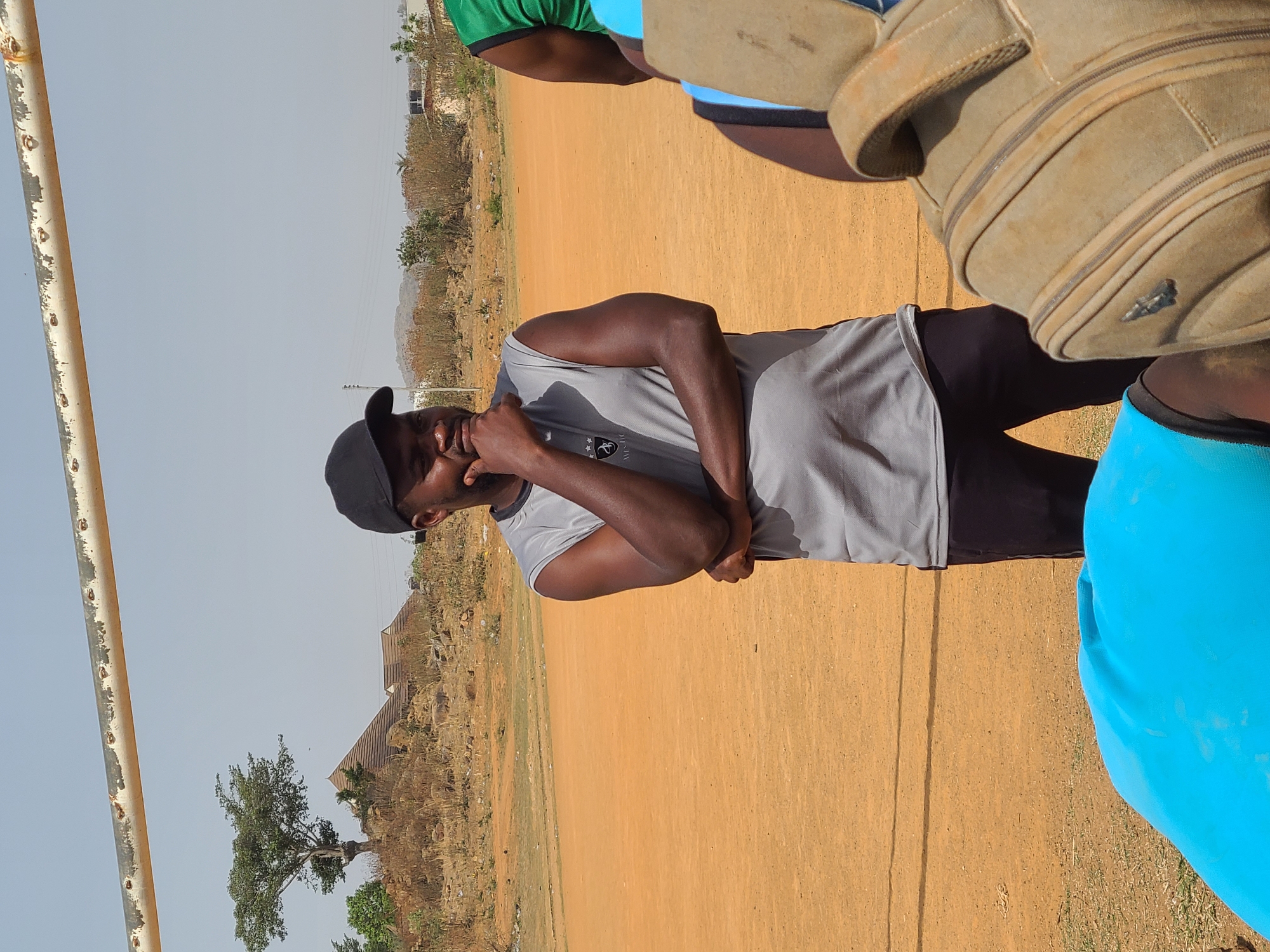 An outdoor scene captured in bright daylight features a dark-skinned man as the central subject. He wears a grey sleeveless shirt with "AMISTAD" and a small emblem visible on the chest, paired with black trousers or shorts and a black baseball cap. He stands with his left hand near his mouth or chin, looking towards the right, while his right arm is bent across his torso, suggesting a thoughtful or casual pose.
Parts of other individuals are visible around him: one person wears a green and black top, another has a bright blue sleeve, and a prominent tan, possibly canvas, backpack with zippers rests on or near one of these partially seen figures.
The setting is an open, reddish-brown dirt field, indicative of dry terrain or an unpaved sports ground. A tall, weathered metal pole, possibly part of a goal post, stands on the far left. The background features sparse, dry vegetation, distant hills or mountains under a clear, pale blue sky, and a few distant structures, including one with a corrugated roof. A lone, leafy tree stands in the mid-ground.
The activity appears to be an informal gathering or break during an outdoor activity, possibly sports-related, given the field and casual attire. The clear sky and bright lighting suggest it is midday or early afternoon. The location cues are consistent with the provided context of Barikin Jirga Summa, Nigeria, hinting at a semi-arid or rural environment.
