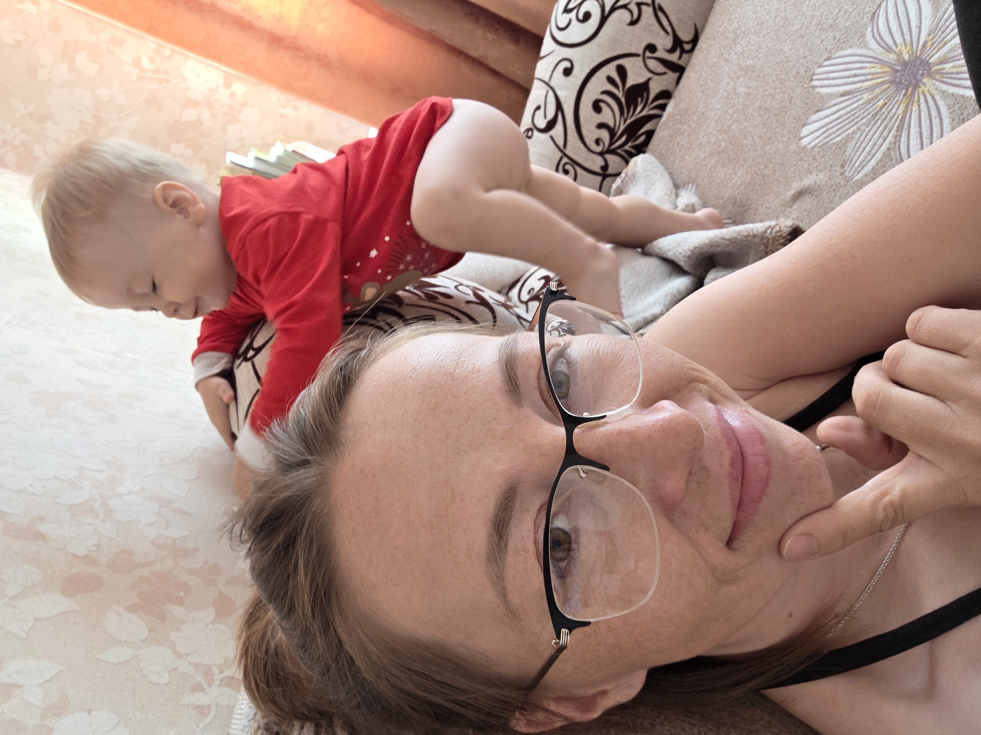 A woman and a baby are on a patterned couch inside a residence, likely in the city of Unknown, Unknown. The woman, wearing glasses and a black tank top, is lying on her back, smiling and touching her chin with her right hand. A silver chain is visible on her chest. The baby, wearing a red, long-sleeved onesie with white star and reindeer patterns, is crawling on top of the woman. A light-colored blanket and patterned cushions are arranged on the couch. The background shows patterned wallpaper and a curtain.