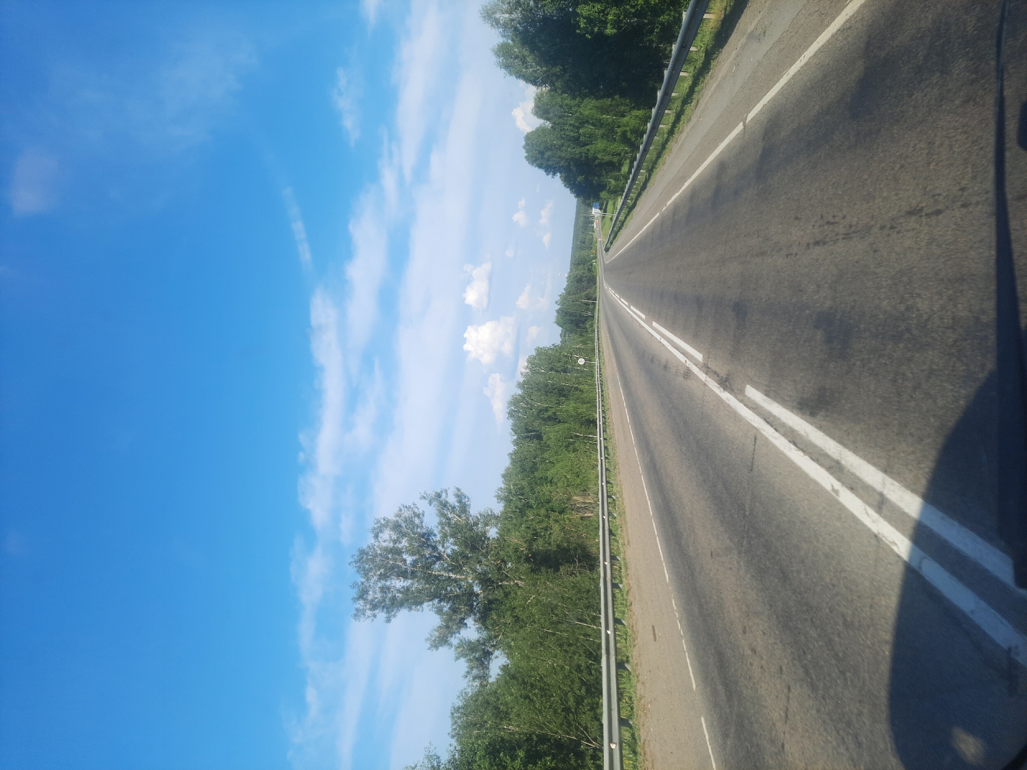 The image depicts a view of a highway in a rural setting, likely in or near Баюновские Ключи, Russia. The perspective is from a moving vehicle, looking forward along the road.
The main subjects are the road itself, flanked by lush green trees on both sides, and a bright blue sky with scattered white clouds. The road is asphalt, with distinct white lines marking the lanes. A metal guardrail runs along the right side of the road. The trees appear to be a mix of deciduous types, with dense foliage suggesting it is daytime during the warmer months.
The activity is travel, as indicated by the road and the perspective. The time of day appears to be morning or afternoon, given the bright sunlight and the clear sky with wispy clouds. The weather seems pleasant and fair. The setting is clearly rural, with the road cutting through a natural landscape dominated by trees. There are no people or specific man-made objects visible, apart from the road, guardrail, and distant, indistinct signs. The overall emotion conveyed is one of serene journeying through nature.