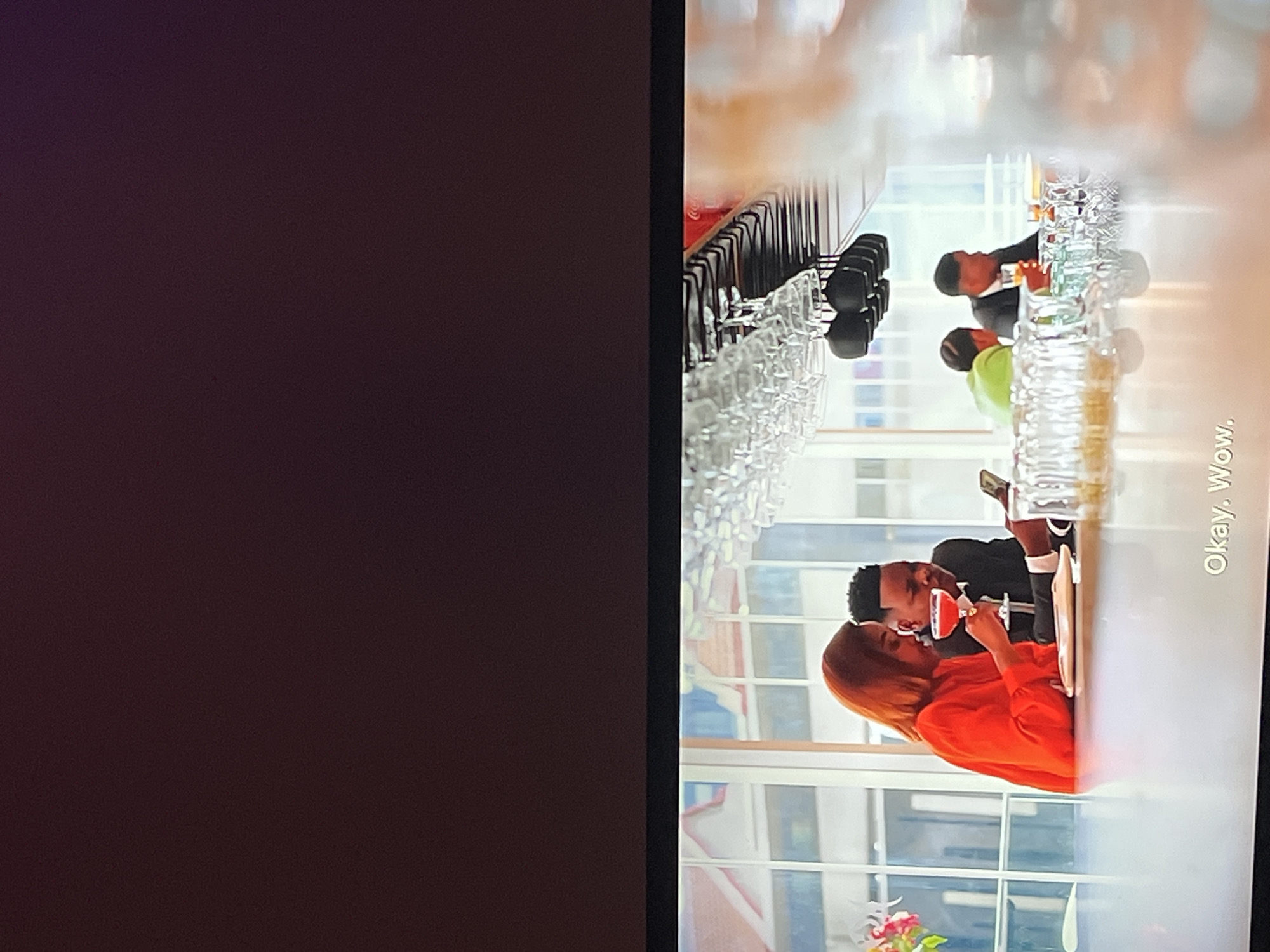 This media file, likely a screenshot or still image from a video, depicts a scene set in a brightly lit interior, possibly a bar or restaurant, with large windows showing a cityscape outside. The image is oriented vertically, with the subjects appearing to be seated at a counter or table.
The primary subjects are a couple. The woman is wearing a vibrant orange top and has long, reddish-brown hair. She is holding a wine glass filled with a red liquid and appears to be interacting with the man beside her. The man is wearing a black suit jacket and a white shirt. He is looking towards the woman and has his hand near her face, suggesting an intimate moment or conversation. Another person, wearing a bright green shirt, is partially visible in the background, also seemingly seated at the counter. In front of the couple and extending into the background are rows of wine glasses, neatly arranged on a rack.
The overall mood seems relaxed and possibly romantic, given the couple's close proximity and the setting. The lighting is bright, suggesting it could be daytime, and the cityscape visible through the windows indicates an urban location. There is a piece of visible text in the lower right corner which reads, "Okay, Wow." The specific location context of Gura Top, Nigeria, is provided but not directly evident from visual cues within the image itself.