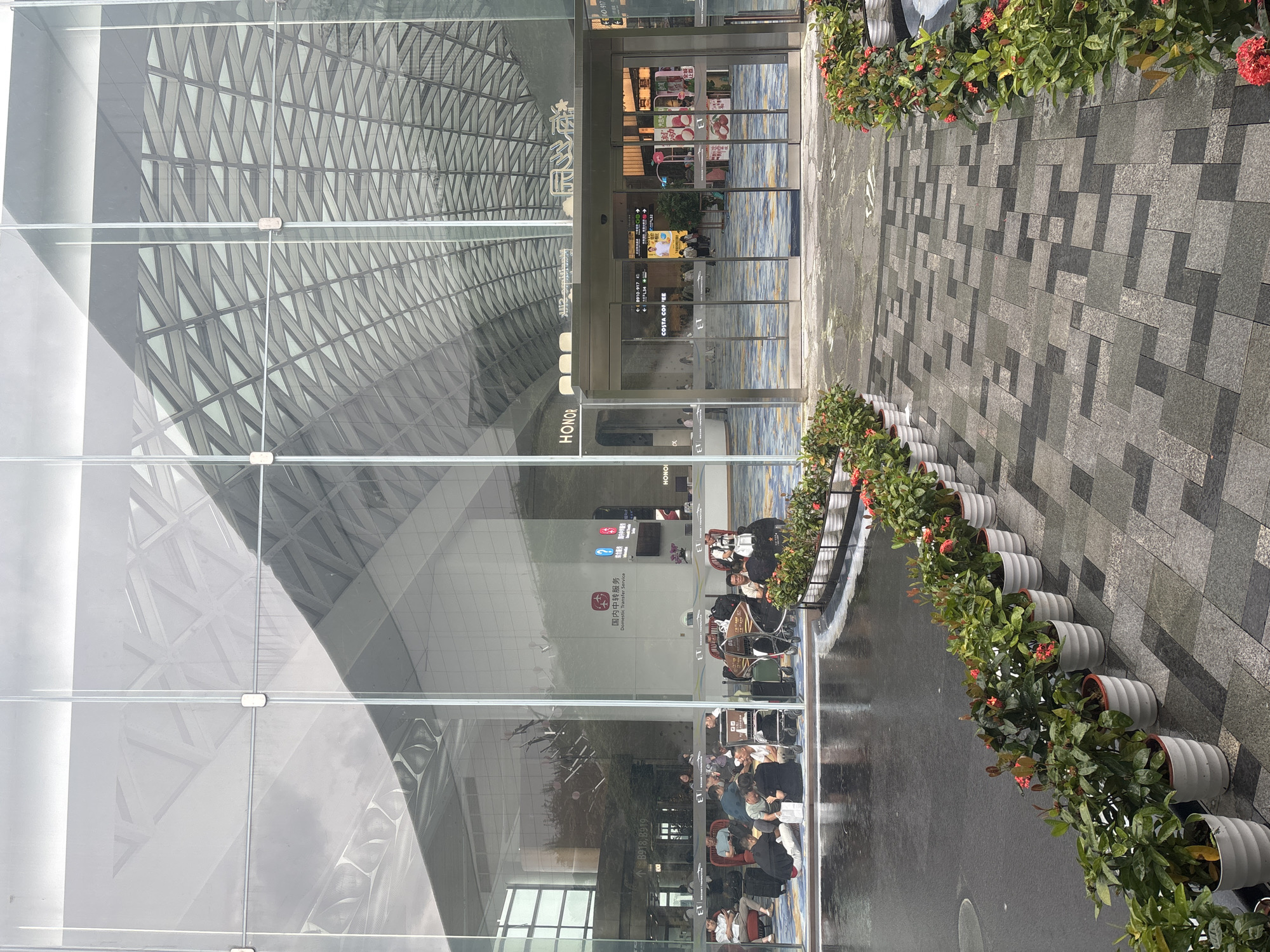 This image depicts the exterior of an airport terminal in Baiyun District, China. The building features a large glass facade with a modern, angular roof structure visible through the reflection. In front of the terminal, a group of people are seated on the ground and in chairs, some with luggage, appearing to be waiting. A row of potted plants with red flowers lines a patterned walkway leading towards the automatic glass doors of the entrance. The ground in front of the building appears wet, possibly from recent rain. Signs in Chinese characters are visible on the building, indicating services like "Domestic Transfer Service."
