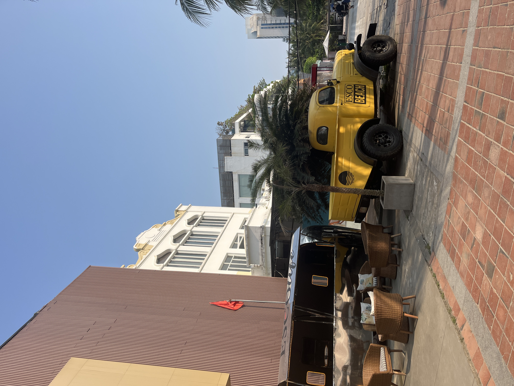 This media file captures an outdoor scene in what appears to be an urban or resort area, likely in Đà Nẵng, Vietnam, under a clear, bright blue sky. The setting suggests a relaxed, possibly beach-adjacent, commercial or leisure space.
The central subjects are two vehicles positioned side-by-side on a paved area. On the right is a vintage-style bright yellow pickup truck, reminiscent of a classic work truck, equipped with large, rugged black wheels. The side of the truck clearly displays the text "ESCO BEACH" in black lettering, along with a stylized wave logo below it. To the left of the yellow truck is a sleek, highly reflective black food truck or trailer, which mirrors parts of the yellow truck and the surrounding environment.
In front of the black trailer, a seating area is arranged with several woven wicker chairs, each featuring a striped cushion. These chairs are set on a concrete path that runs parallel to a brick-patterned sidewalk or plaza area.
The background features various architectural elements. On the far left, there is a building with a textured, reddish-brown corrugated facade, from which a small red, flag-like object extends. Adjacent to it stands a multi-story white building with ornate, classical-inspired detailing along its roofline and windows. Further back, more modern white buildings with flat roofs and large windows are visible, interspersed with lush palm trees. In the distance, a taller, contemporary building rises above the other structures.
The strong, distinct shadows indicate bright sunlight, suggesting it is midday or early afternoon. The overall atmosphere is sunny and inviting, consistent with a pleasant day for outdoor activities or dining. No people are actively visible, but the setup implies a space designed for public use.