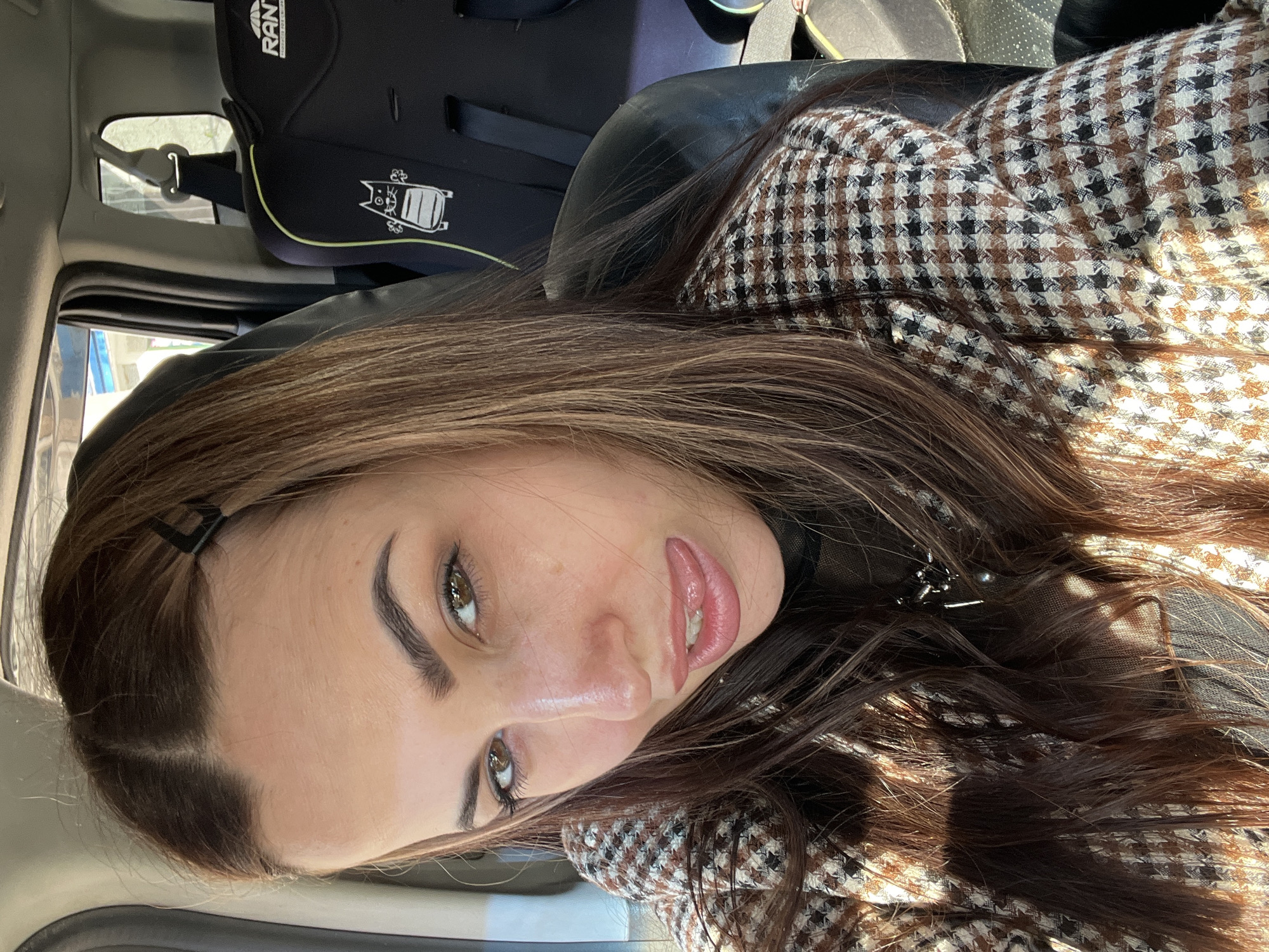 The photograph shows a young woman in the front seat of a vehicle. She is facing the camera, and her head is tilted slightly. Her hair is long and brown, with a black hairpin visible near her temple. She is wearing a brown and white checkered coat. The background shows the interior of the car, including a black car seat. The location is unknown.