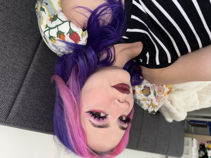 An indoor, eye-level shot captures a female individual reclining on a bed. Her head is positioned to the right, with her body angled slightly left towards the camera, maintaining direct eye contact. She has long, wavy hair, distinctly colored in alternating sections of bright pink and vibrant purple. Her face is heavily made up, featuring prominent winged black eyeliner, bright pink eyeshadow, rosy blush, and dark plum-colored lipstick. She is wearing a black and white horizontally striped, ribbed tank top or camisole. Her left arm is visible on the left side of the frame. She is supported by a floral patterned pillow with red, orange, green, and blue motifs on a light background, and a grey upholstered headboard is visible behind her and to her right. A fluffy white blanket or throw is partially visible on the bed to her left. The scene is consistent with a domestic bedroom setting in the City of Zagreb, Croatia.