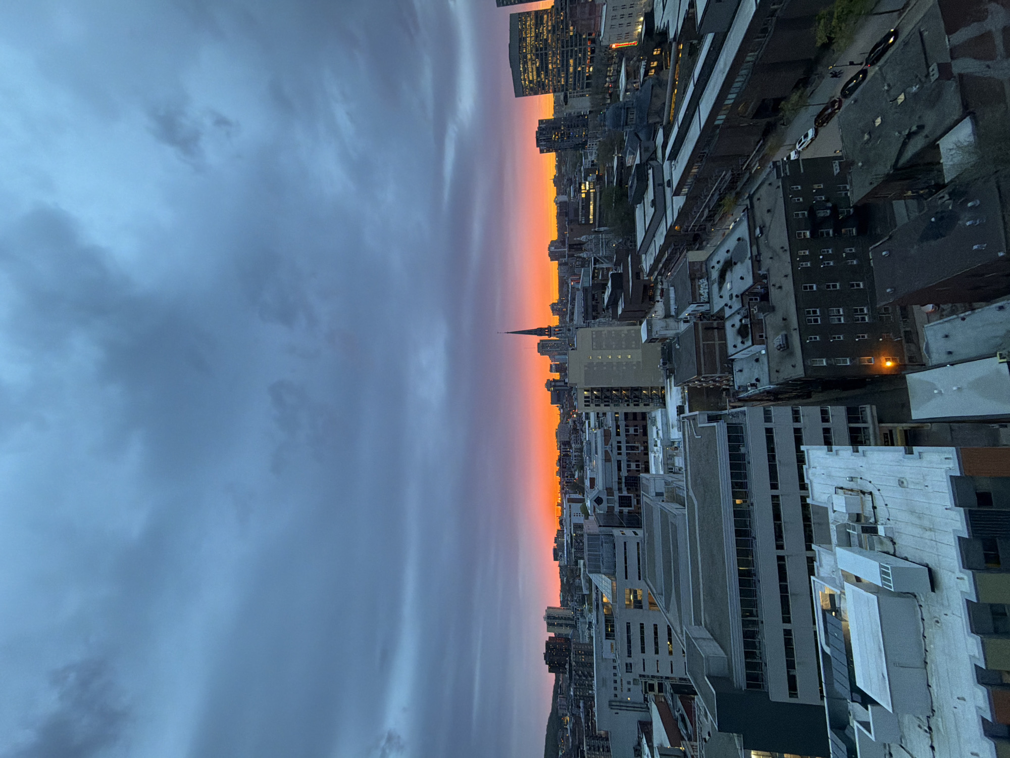 From an elevated perspective in Montreal, Canada, the image captures a cityscape under a dusk sky. The sky transitions from grey clouds on the left to an orange and pink horizon in the center, indicative of a sunset. This coloration casts a warm glow on the buildings below. The architectural structures range in height and style, with visible windows indicating occupancy. Roads and cars are discernible between the buildings. In the middle ground, a spire-like structure stands prominently, possibly a church or religious building.