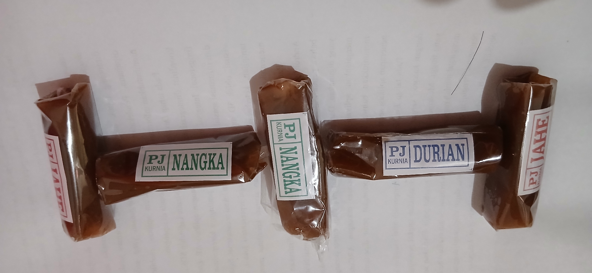 The image shows a stack of four individually wrapped food items. Each item appears to be a cylindrical piece of confectionery or fruit leather. Three of the items have labels with the text "PJ NANGKA", "PJ DURIAN", and "PJ JAHE", with smaller text "KURNIA" above "PJ". One item at the top is partially visible and appears to have a label with red text that reads "PJ TATE". The items are arranged vertically, with two items at the bottom, one above them, and one at the very top. The background is a white surface with faint text printed on it, resembling paper. There is no indication of the city of Samarinda, Indonesia, or any specific location within it in the image.