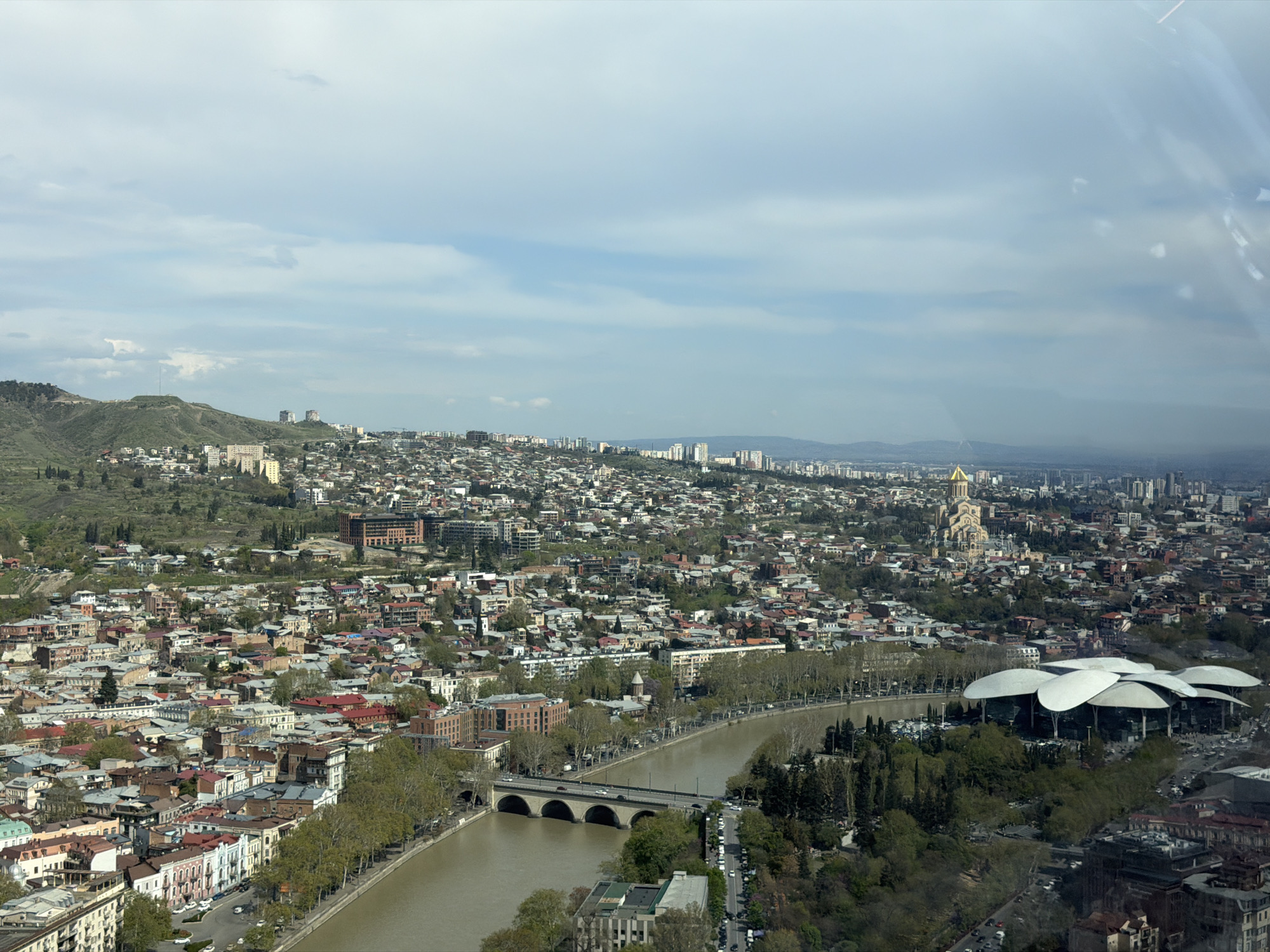 This high-angle, panoramic shot provides an expansive view of a city, identified as Tbilisi, Georgia, under a partly cloudy sky. The urban landscape stretches across a wide area, flanked by green, rolling hills and distant mountains that fade into a light haze.
The Kura River (Mtkvari River) flows prominently through the heart of the city, its brownish water reflecting the overcast conditions. Several bridges span the river, with one multi-arched stone bridge clearly visible, accommodating vehicular traffic. Both sides of the river are lined with trees, creating green corridors within the dense urban fabric.
The city is characterized by a mix of architectural styles, from older, tightly packed residential buildings with varied roof colors to more modern, taller structures. Two prominent landmarks stand out: the majestic Holy Trinity Cathedral of Tbilisi (Sameba Cathedral), with its distinctive golden dome, is visible on a hill on the right side of the frame. Closer to the river, on the right, is the modern Public Service Hall, recognizable by its unique, white, mushroom-like roof design.
The time of day appears to be daytime, likely late morning or early afternoon, with soft, diffused lighting from the overcast sky. Cars are visible on roads and bridges, indicating ongoing urban activity, though no specific human figures or legible text are discernible due to the distance. The scene captures a bustling city nestled within a natural, hilly environment.