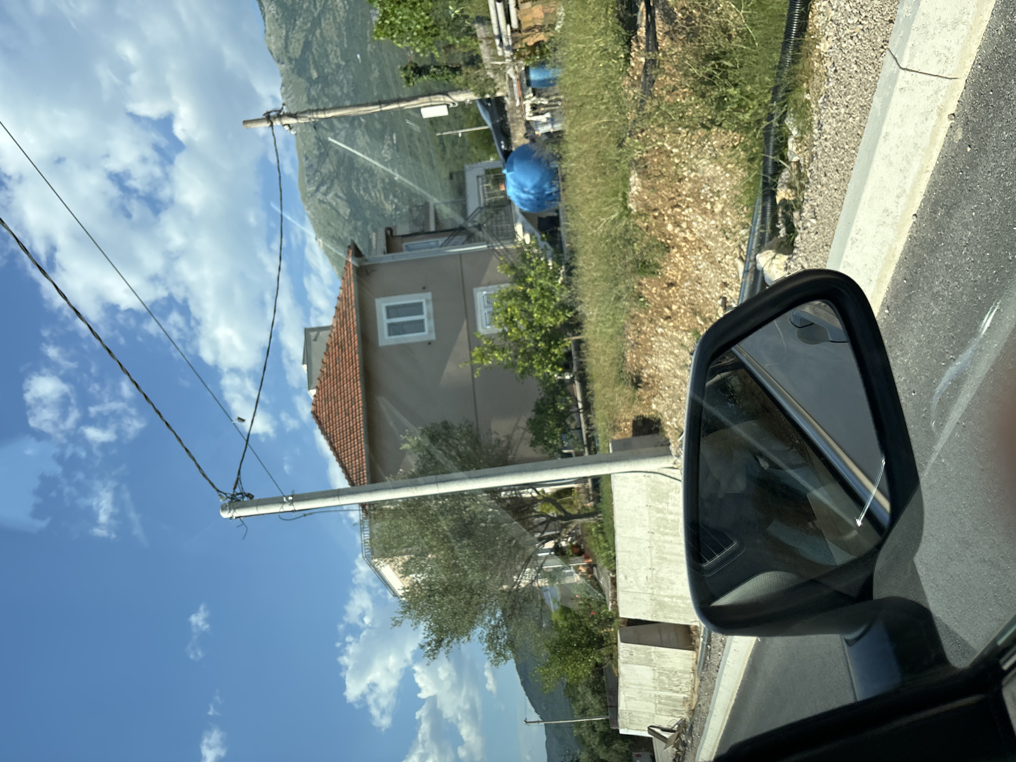 This is a vertical, low-angle, outdoor, daytime shot of a residential area in Radanovići, Montenegro. The image is captured from the perspective of someone in a car, looking out the side window and towards the side-view mirror.
The scene depicts a house with a terracotta roof, beige walls, and white-framed windows. Behind the house, a steep, green mountain slopes upwards, partially obscured by trees. In the foreground, a white power pole with black wires stretches across the frame, connecting to a building on the right. Adjacent to the road, there is a grey concrete curb and a gravel area with dry grass and some green shrubbery.
The side-view mirror reflects the interior of the car, including what appears to be a person in the passenger seat wearing a white garment and a blue item. Outside the mirror's reflection, a bright blue tarp is visible, possibly covering something in the yard. The sky is a vibrant blue with scattered white clouds, suggesting a sunny day.
The overall impression is of a quiet, rural, or semi-rural setting in the Mediterranean. The lighting indicates it is likely late morning or early afternoon. There is no visible text in the image.