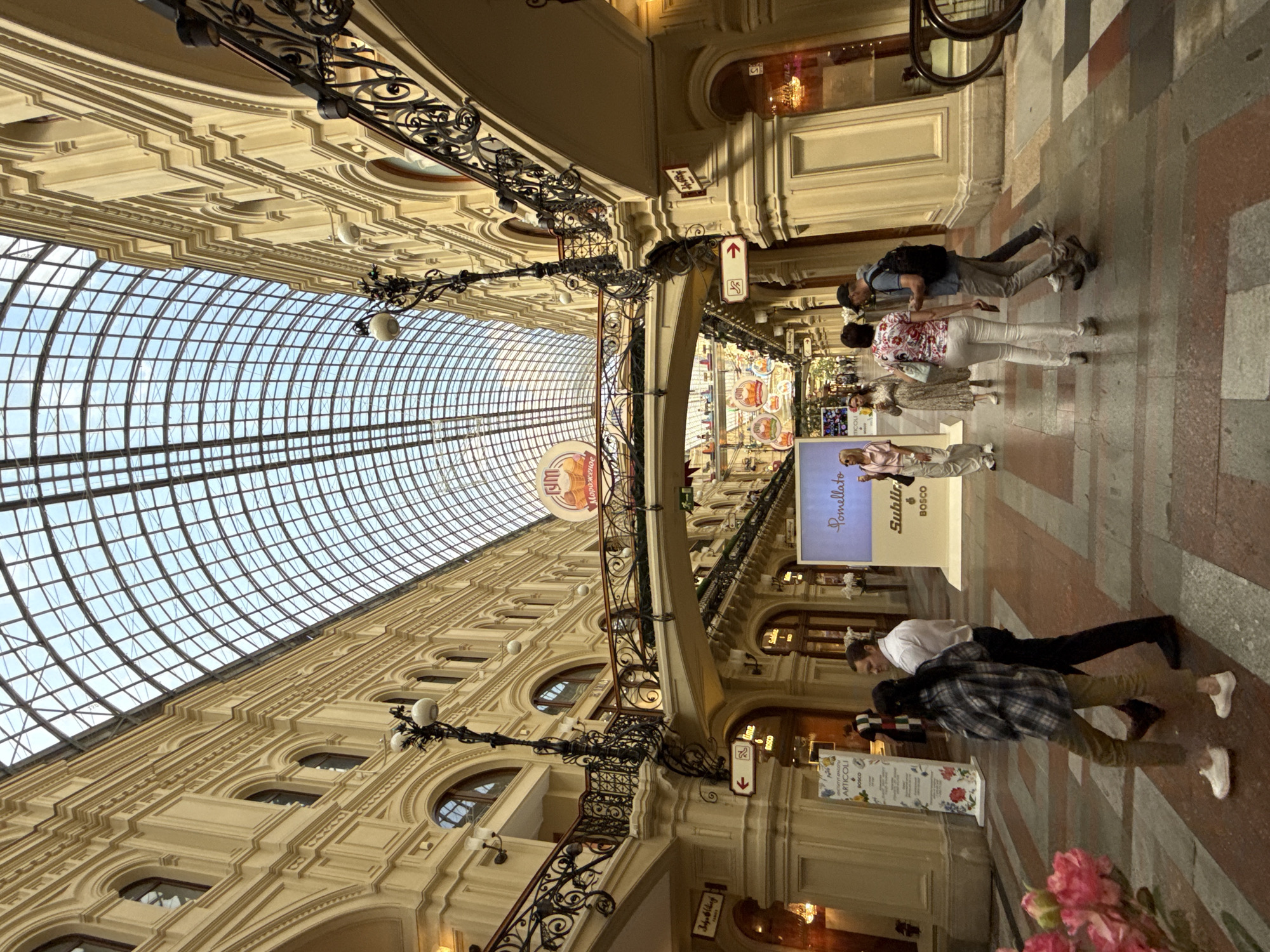 The image depicts an interior view of a large, covered shopping arcade with a distinctive arched glass roof, typical of the GUM department store on Red Square in Moscow, Russia. The arcade's architecture features ornate yellow facades with multiple levels of balconies and intricate black metal railings. Several decorative street lamps are mounted on the walls.
In the foreground, a group of people are walking on a patterned floor made of large, rectangular tiles in various shades of red, gray, and beige. On the left, a person in a plaid shirt and olive pants walks away from the camera, followed by a man in a white shirt and dark pants. Further down the arcade, a woman in a floral shirt and white pants walks towards the camera, accompanied by a man in a blue shirt and gray pants, who is looking at his phone. Other individuals are visible in the background, some browsing shop displays, others walking along the arcade.
Above the shoppers, a prominent bridge spans the width of the arcade, adorned with decorative railings and signs. One sign features the Russian word "ГУМ" (GUM) with an image of ice cream, suggesting a food-related advertisement. Another sign displays "Pomellato" and "Sublime BOSCO". Numerous shop entrances are visible on either side of the arcade, displaying merchandise and illuminated interiors. The overall scene conveys a bustling atmosphere of a high-end shopping environment.