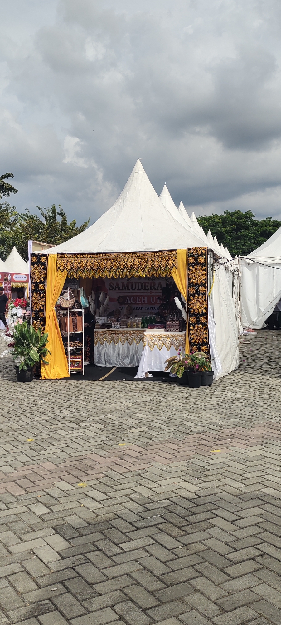 The image shows an outdoor market stall in Meunasahaluedrienlhoksukon Barat, Indonesia, under a cloudy sky. The stall is set up under a white conical tent with decorative gold and black fabric draped around its opening. Inside the stall, there are tables displaying various items, including what appear to be food products in jars and bottles, as well as woven baskets and bags. A sign above the stall reads "SAMUDERA ACEH UT". To the left of the main stall, another white tent is visible, with a display of artificial flowers. Plants in black pots are placed around the main stall. The ground is paved with interlocking brick pavers. In the background, trees and more tents are visible, suggesting a larger event or market.