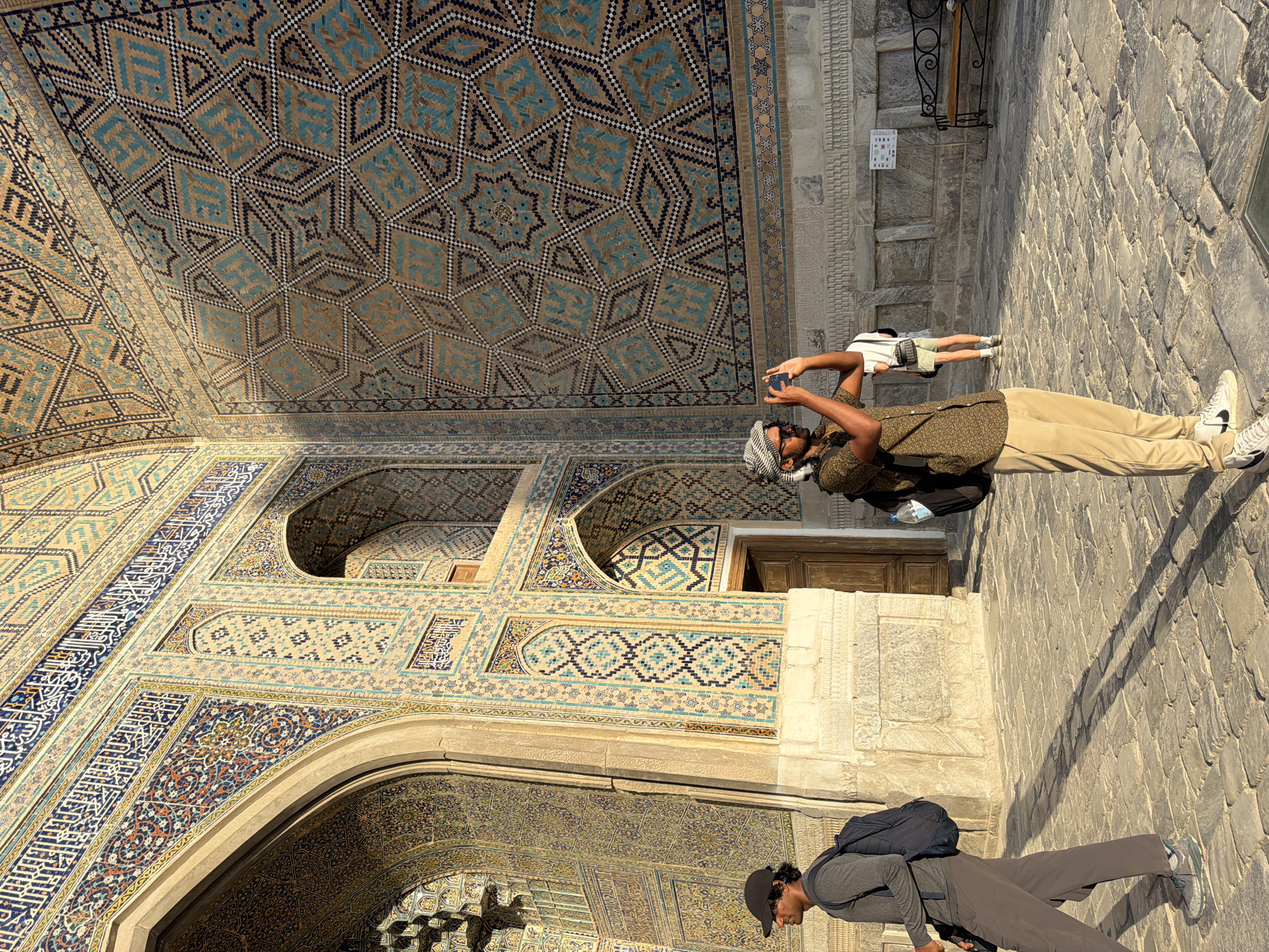 A man wearing a grey long-sleeved shirt, grey pants, and a dark backpack is walking to the left. In the foreground, a man with a patterned short-sleeved shirt, khaki pants, and a headscarf is taking a picture with his phone. Behind him, a woman in a white t-shirt and shorts is visible, as is a bench. The background features the intricate tilework of the Registan complex in Samarkand, Uzbekistan, illuminated by the sun.