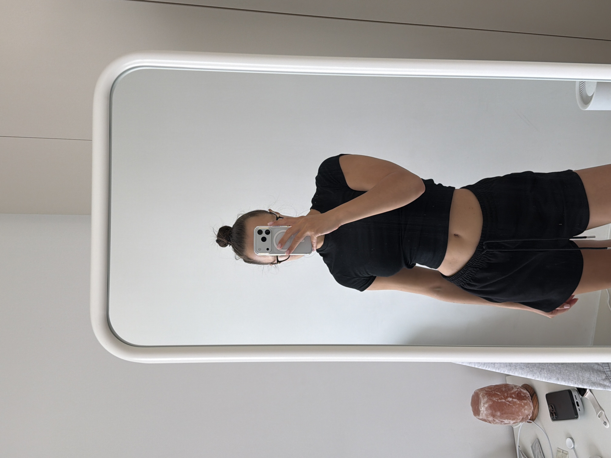 A woman takes a mirror selfie. She is wearing a black top and black shorts. She has her hair in a bun and wears glasses. A phone covers her face in the mirror. A nightstand is beneath the mirror, on which sits a salt lamp, a phone, and a few small electronics. The room is white. I cannot determine the location in Belgrade, Serbia from this image.