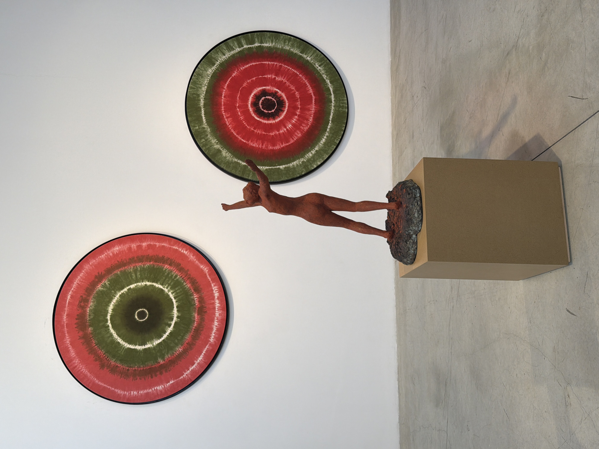 The image depicts an art installation in a gallery. On the white wall, two circular artworks with concentric rings of red and green are displayed. In the foreground, a reddish-brown statue of a female figure with arms outstretched upwards stands on a dark, rocky base, which is placed on a light brown cubic pedestal. The floor is a grey concrete surface. The scene appears to be set in a modern gallery space, potentially in Tel-Aviv, Israel, given the context of the request.