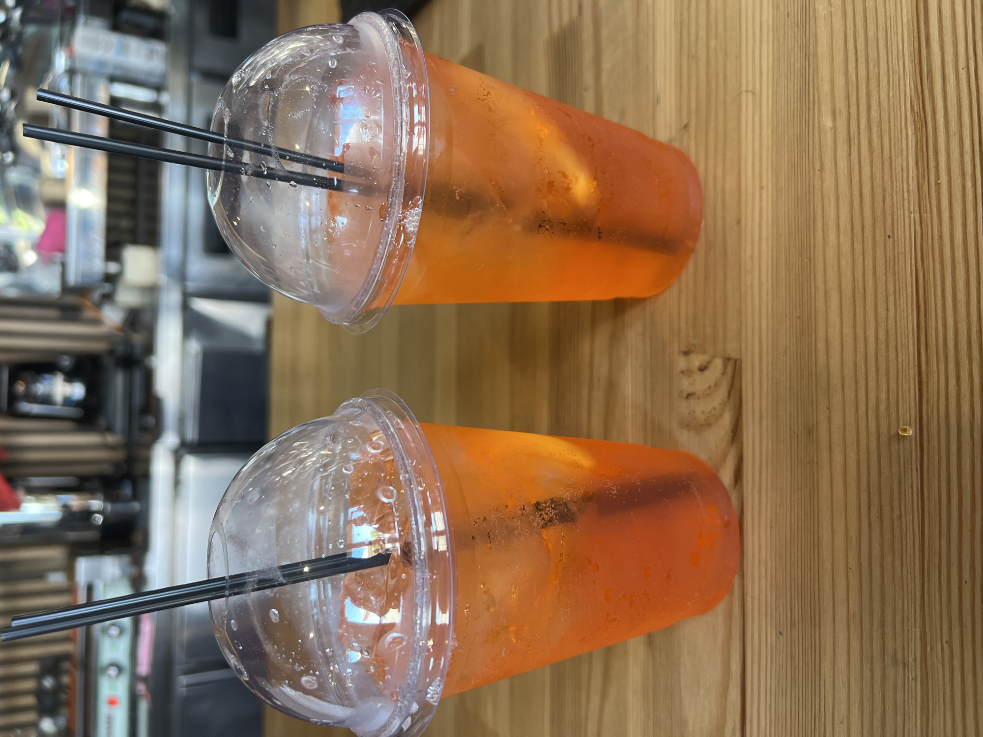 Two plastic cups filled with orange beverages and ice sit on a wooden table. Black straws are visible in each cup. The background is blurred, showing a bar or restaurant setting with kitchen equipment and patrons. The location is identified as Fontanka, Ukraine.