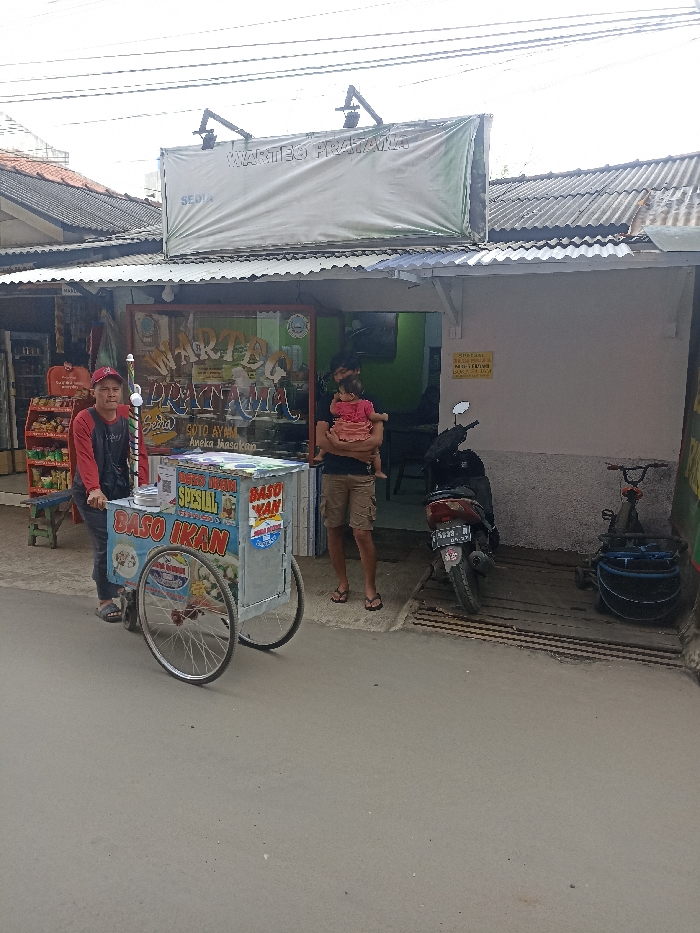 The image shows a street scene in Jatinangor, Indonesia, at coordinates -6.934 latitude and 107.775 longitude. A man is standing next to a food cart labeled "BASO IKAN," which is parked in front of a storefront. The storefront appears to be a restaurant or food stall, with a sign above it that reads "WARTEL PRATAMA" in Indonesian. The man is wearing a red shirt, black pants, and a blue cap. He is holding a red and white striped umbrella and appears to be preparing to move the cart. To the right of the cart, a man in a green shirt is carrying a young girl in a pink dress. The man is facing the camera and looking at the cart. Behind them is a black motorcycle parked in front of the storefront. The storefront has a large window that displays various items including a sign that says "SOTO AYAM." A second motorcycle is parked at the edge of the sidewalk. The scene is typical of a busy street in a small town. The pavement is dusty and there are shadows from buildings and trees. The weather appears to be overcast and the sky is white.