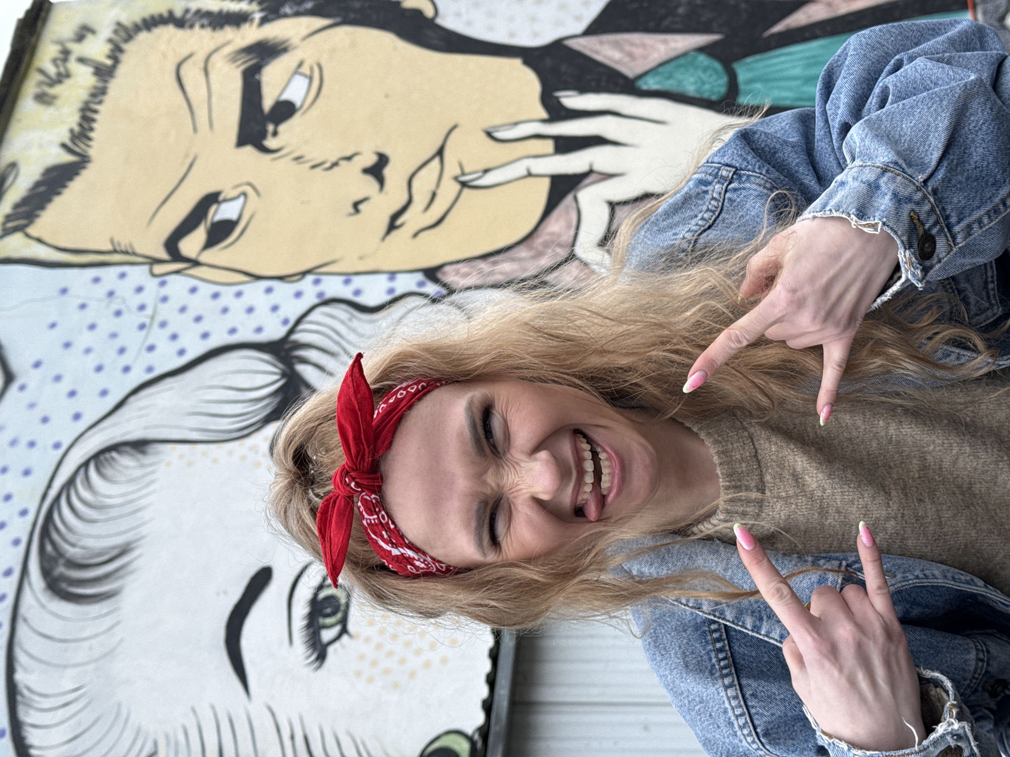 A blonde woman with a red bandana on her head is smiling and sticking her tongue out, making a "rock on" gesture with both hands. She is wearing a denim jacket over a beige sweater. Behind her is a piece of street art featuring cartoon-like faces. The location is Chaikovsky, Russia.