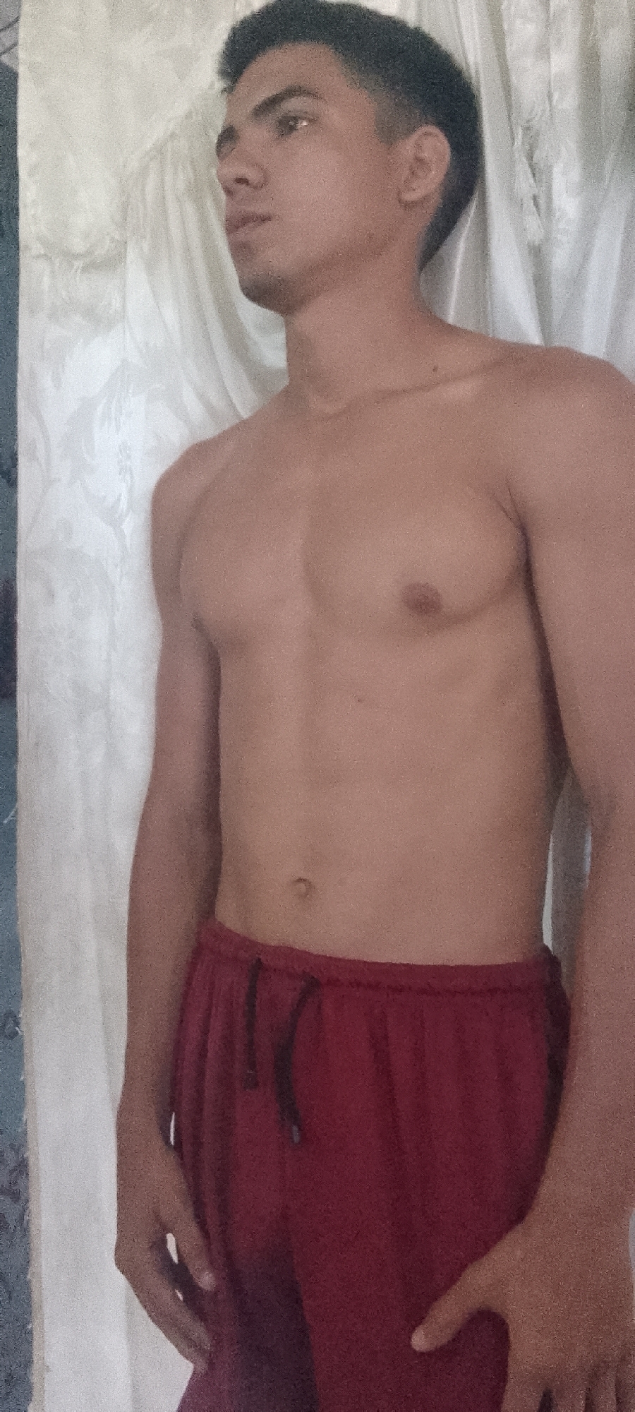 A young adult male, with dark, short hair and medium skin tone, is depicted standing indoors. He faces partially to the right, looking upwards and to the right. The individual is shirtless, revealing his upper torso and abdomen, and wears dark red athletic shorts with a visible black drawstring. His right hand is positioned at his waist, resting on the shorts. The background consists primarily of a light-colored, patterned fabric, consistent with a curtain or tapestry, covering most of the vertical space behind him. A darker, possibly blue or grey, textured surface is partially visible on the far left edge of the image. No discernible landmarks or external features are present to identify a specific location within the city of Juan Griego, Venezuela; the setting appears to be a generic interior space.