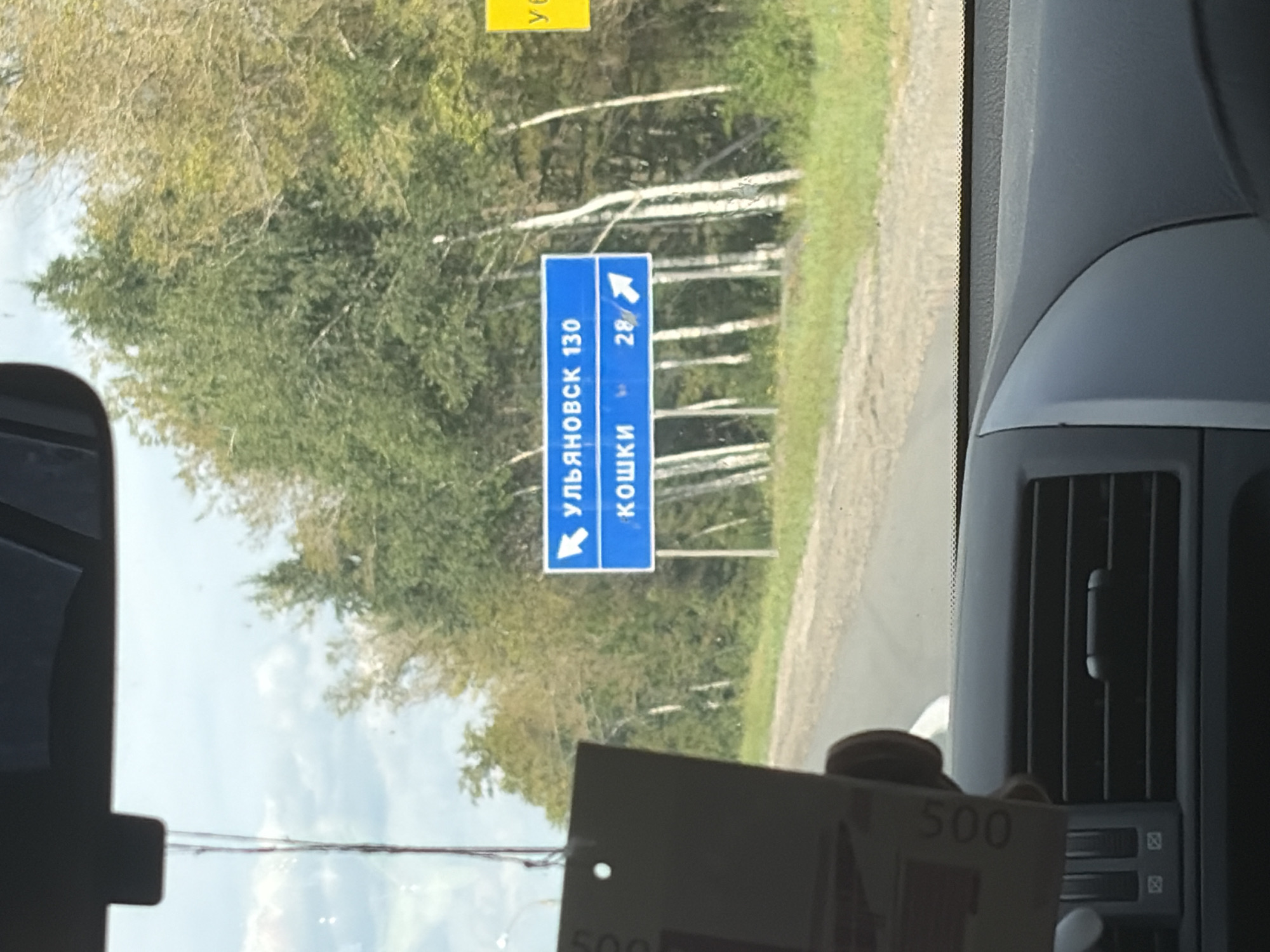 A blue road sign is visible in the image, indicating directions to "ULYANOVSK 130" and "KOSHKI 28". The sign is positioned on the right side of a paved road, with trees and greenery in the background. A yellow sign is also partially visible to the right of the blue sign. In the foreground, a car's dashboard and air vent are visible, suggesting the photo was taken from inside a vehicle. A dark object with the number "500" printed on it is hanging from the rearview mirror. The scene appears to be in a rural or semi-rural setting.