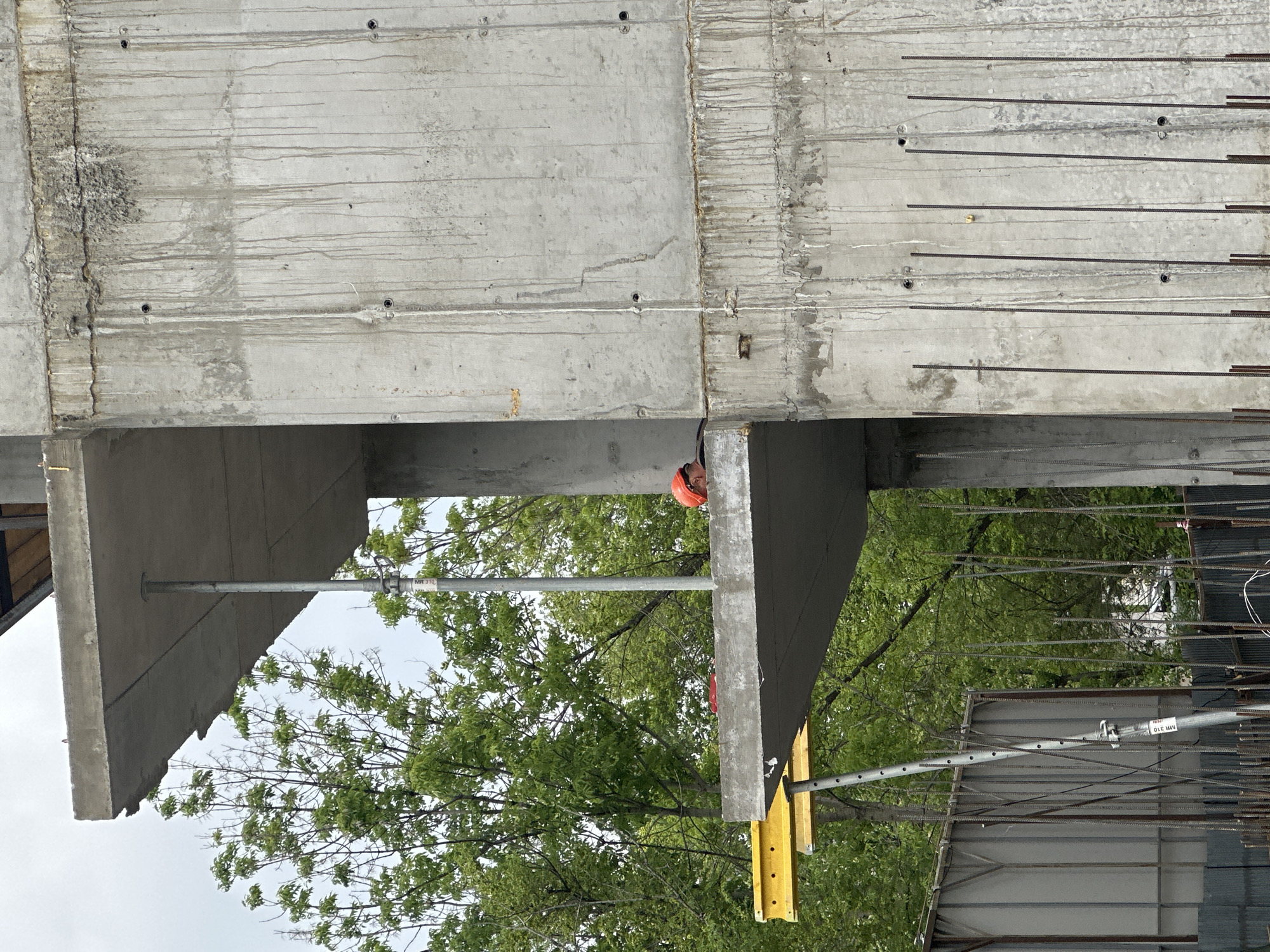 The image shows a construction site in Khabarovsk, Russia. A partially completed concrete structure dominates the scene, exhibiting unfinished surfaces, exposed rebar, and support beams. A construction worker wearing an orange hard hat is visible between the concrete structures. Green trees fill the background, and metal scaffolding is present in the lower right of the frame.