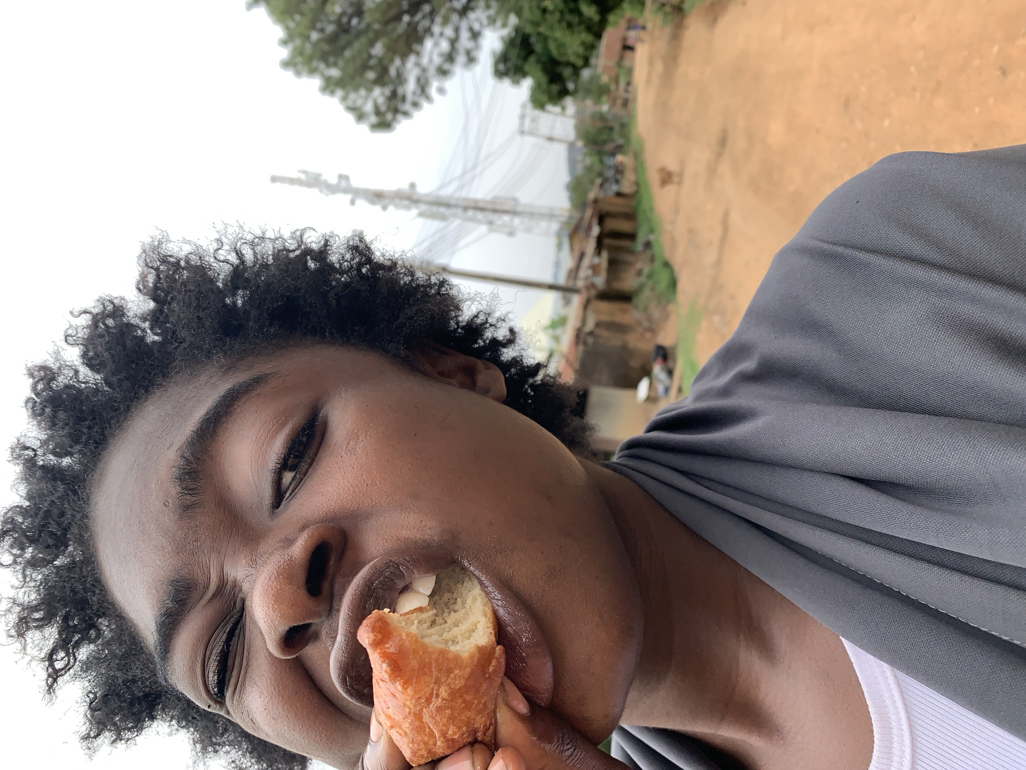 A close-up shot depicts a person with dark skin and curly dark hair, holding a piece of fried dough (likely a type of local pastry) to their mouth. The person's eyes are squinted, and they appear to be taking a bite. The individual is wearing a white undershirt and a gray, possibly linen, garment draped over their shoulders. In the background, there is a dusty, unpaved road with sparse vegetation, and a utility pole supporting a network of wires. The location is indicated as Jos, Nigeria based on the surrounding environment of the city.