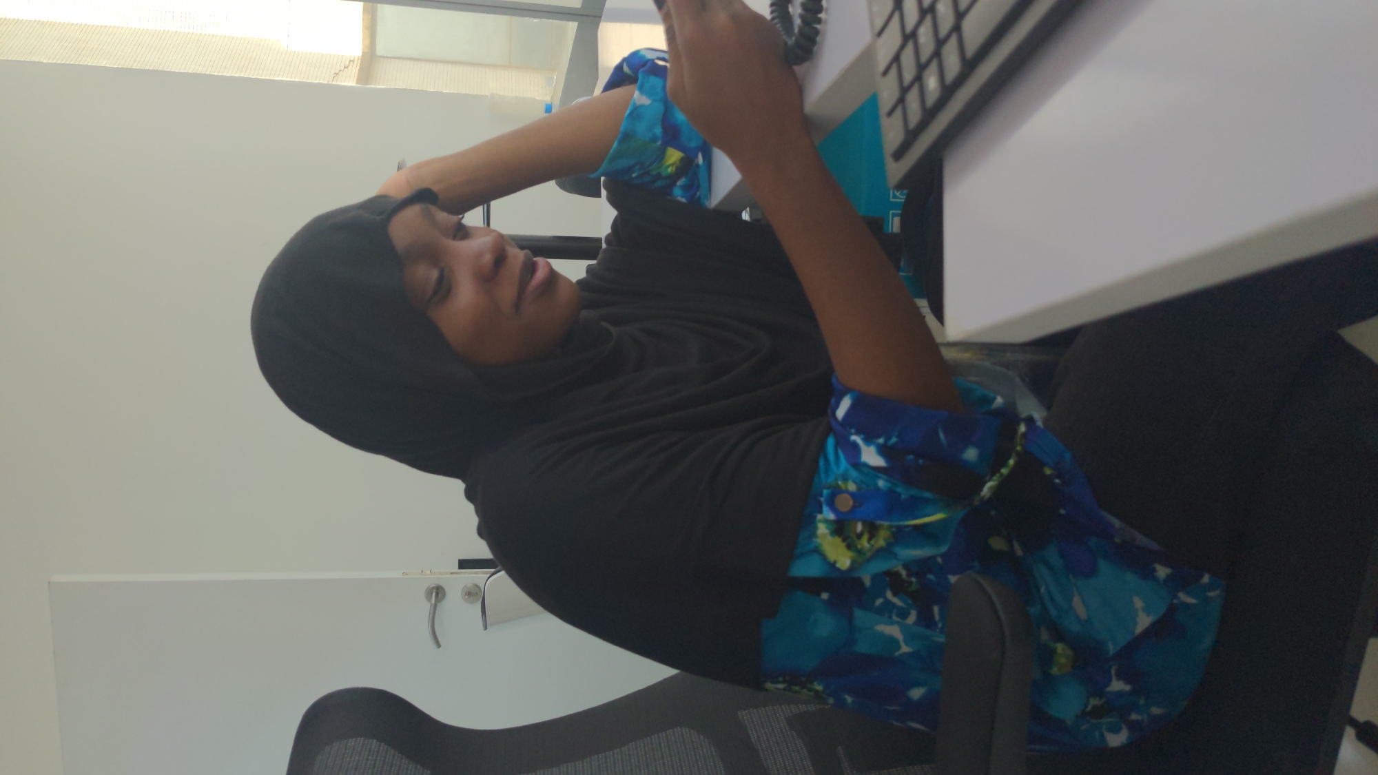 A young adult female is depicted in a brightly lit indoor office setting, likely in Abuja, Nigeria. She is seated on a grey and black office chair at a white desk, engaged with a device held in her hands, which appears to be a telephone receiver with a coiled cord, or possibly a smartphone. She wears a black hijab covering her head and neck, along with a black long-sleesleeved top. Her sleeves feature a distinctive and vibrant pattern of blue, white, and hints of green, resembling abstract or floral motifs.
Her head is slightly tilted, and her gaze is directed downwards towards the object in her hands, suggesting focus or contemplation. A faint, gentle expression is visible on her face. A black computer keyboard is partially visible on the white desk to her right. The background features a plain white wall, with a white cabinet or storage unit with silver handles visible behind her to the left. To the top left, a window or light source allows natural light to enter, illuminating a textured exterior structure. The scene portrays a calm and professional atmosphere during what appears to be daytime. No discernible text is visible in the image.