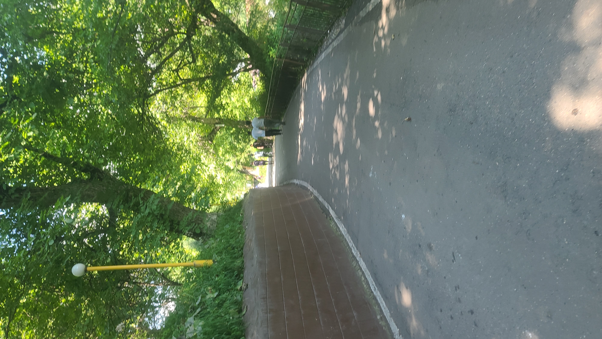 A paved pathway curves through a park in Arstanbap, Kyrgyzstan, shaded by dense green trees. A retaining wall with horizontal brown planks lines the left side of the path. A yellow lamppost with a white globe is visible on the left, near the top of the retaining wall. In the distance, several figures walk along the path. The ground is dappled with sunlight and shadows. A metal fence runs along the right side of the pathway.