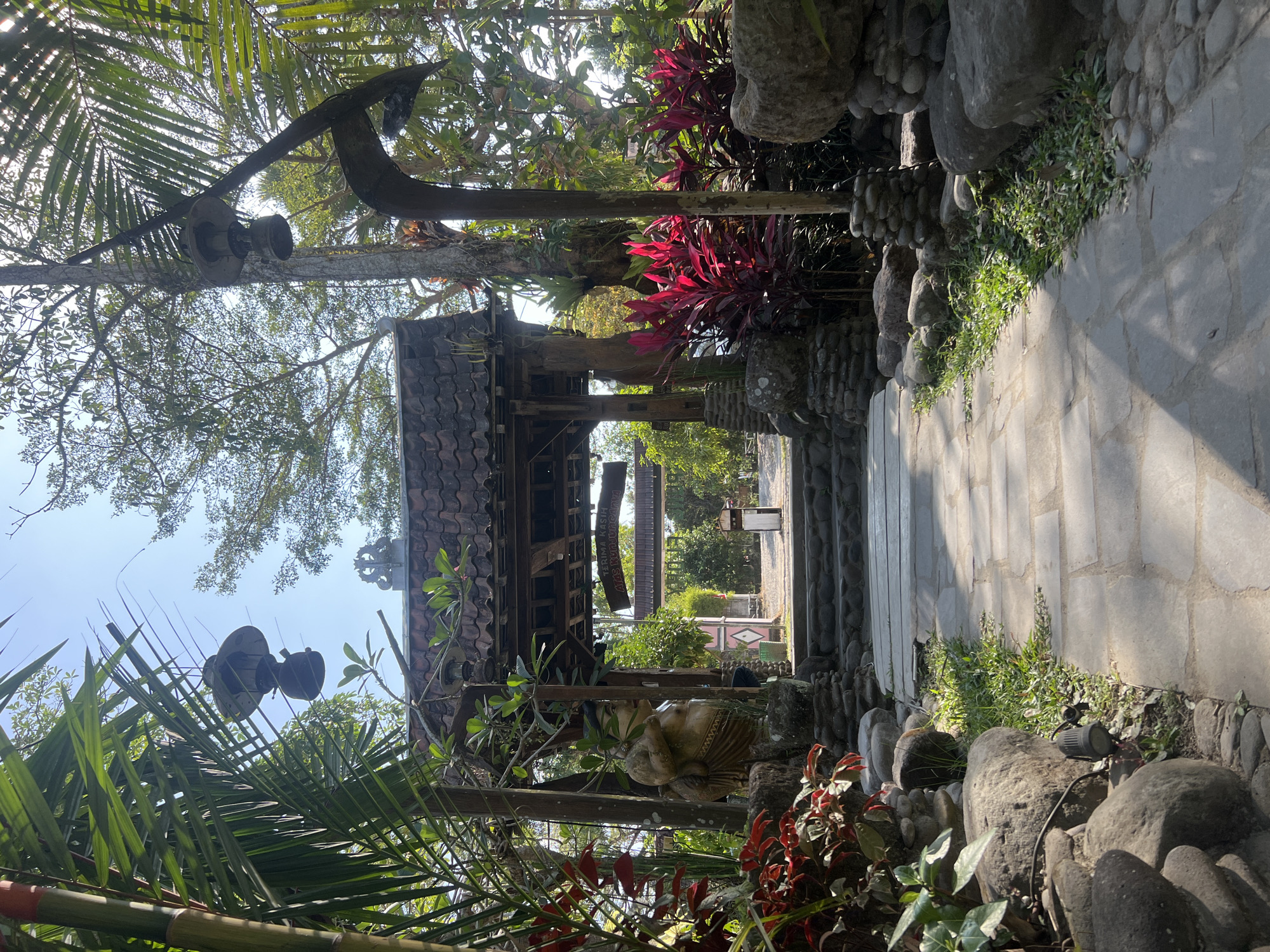 A sun-dappled stone pathway leads through a lush, tropical garden towards an ornate wooden gate. The gate, adorned with a tiled roof and intricate carvings, marks the entrance to a complex that appears to be a cultural attraction or a resort. Two traditional Balinese statues, one of a stout, smiling figure, flank the pathway leading up to the gate. Vibrant red foliage and towering palm trees frame the scene, creating a sense of tranquility and exoticism. Overhead, traditional oil lamps hang from branches, adding to the ambiance. A sign above the gate reads "TERIMA KASIH ATAS KUNJUNGANNYA," which translates to "Thank you for your visit" in Indonesian, indicating this is a visitor destination in Tlogoadi, Indonesia.