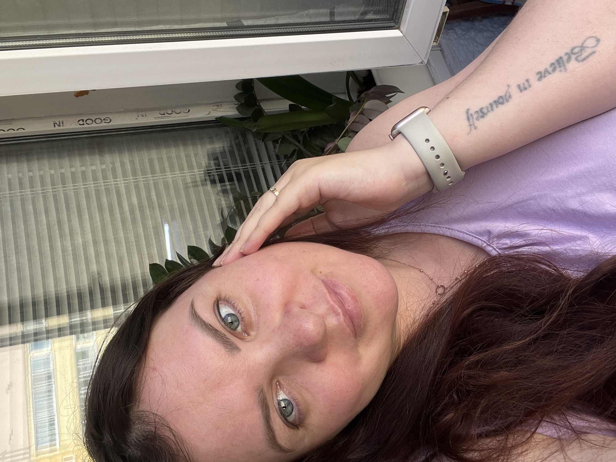 A woman with long brown hair and blue eyes is taking a selfie in what appears to be an indoor setting. She's wearing a lilac-colored top and has a light-colored smartwatch on her left wrist. A tattoo is visible on her left forearm. She's touching her forehead with her hand. The background includes a window with blinds, and part of a building is visible outside, along with what seems to be a potted plant on the window sill. The window frame has the name "GOOD IN" printed. The specific location in Noviy, Russia cannot be determined from the image alone.