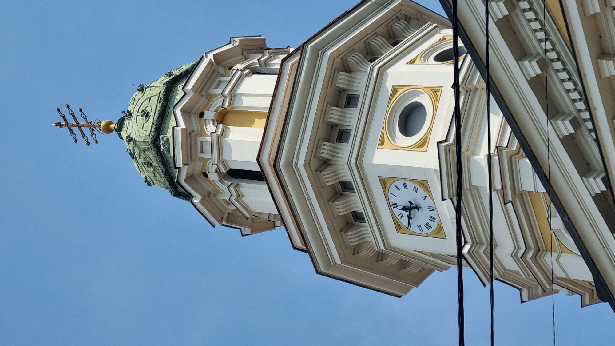 This image captures the upper section of a white church tower with a green dome, topped by a golden cross, against a clear blue sky. A clock face with Roman numerals is visible on one side of the tower. The architectural style suggests it is an Orthodox church. The scene is likely located in Uzhhorod, Ukraine, as indicated by the architectural style commonly found in the region. The foreground shows dark electrical wires crossing horizontally.