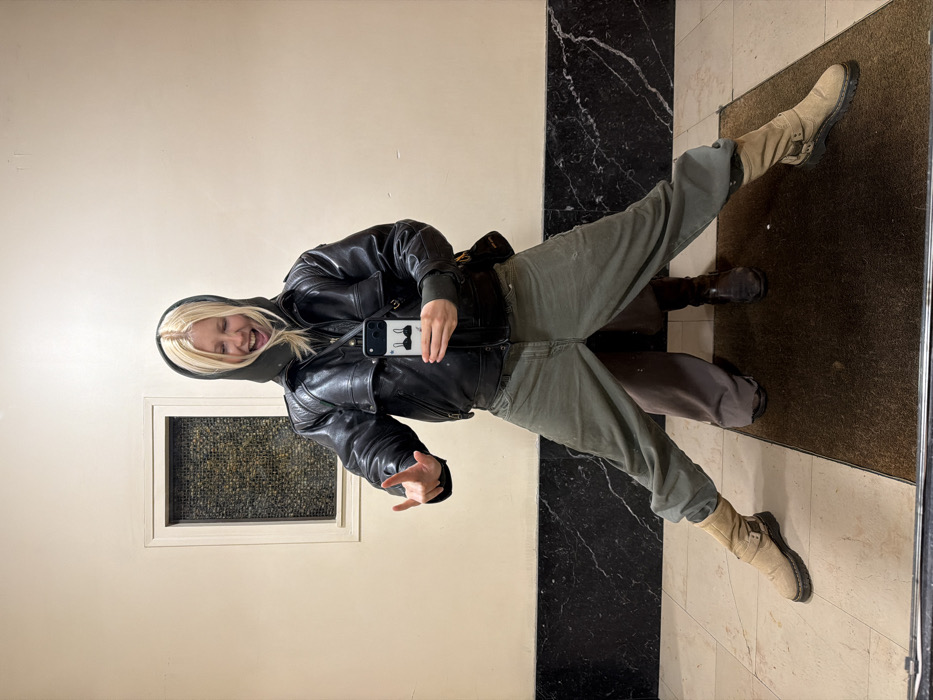 A person takes a mirror selfie. The individual wears a black leather jacket, green pants, and light-colored boots. They also sport a grey hooded sweater. A marble-patterned wall and a white wall are visible in the background. The person is making a hand gesture while sticking their tongue out. The smartphone case shows an image of a cat's whiskers and bow tie.
