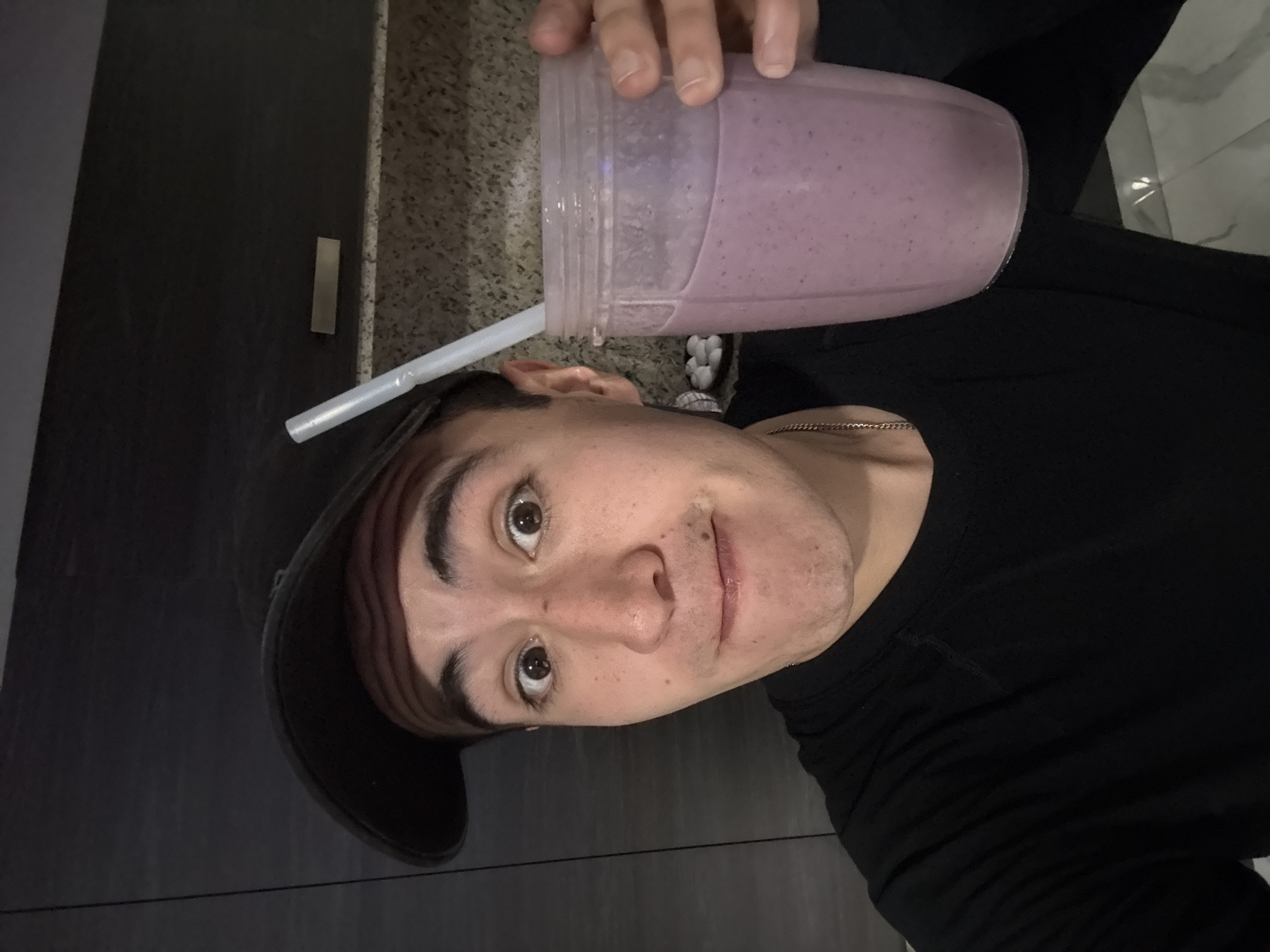 A man wearing a black shirt and a black baseball cap is holding a clear plastic cup filled with a purple smoothie and a white straw. He has light skin, dark eyebrows, and brown eyes. He is indoors; the background shows a dark gray cabinet or wall to his left and a light-colored countertop to his right. The scene appears to be lit with artificial light. There is no verifiable information present in the image to confirm that the location is San Bernardino Tlaxcalancingo, Mexico.