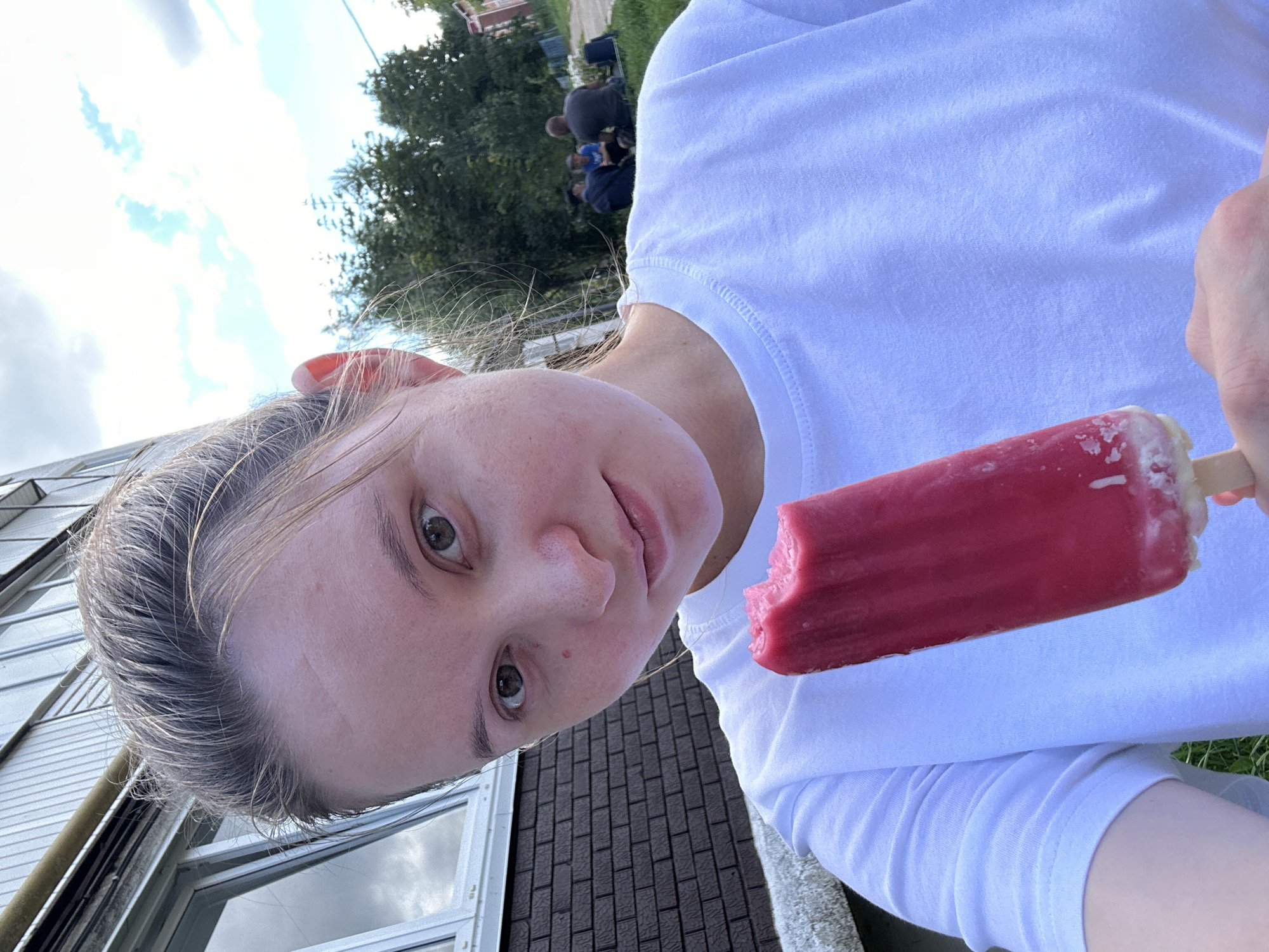 A young woman, with blonde hair pulled back and a light complexion, is taking a selfie outdoors. She is wearing a white t-shirt and holding a partially eaten pink popsicle in her right hand. Her gaze is directed at the camera. In the background, behind her, a building with a brick lower wall and a metallic upper section is visible, along with greenery and trees. Further in the distance, three individuals are seated on the grass, their backs to the camera, suggesting a communal outdoor setting. The sky above is partly cloudy. The location is identified as Noviy, Russia.