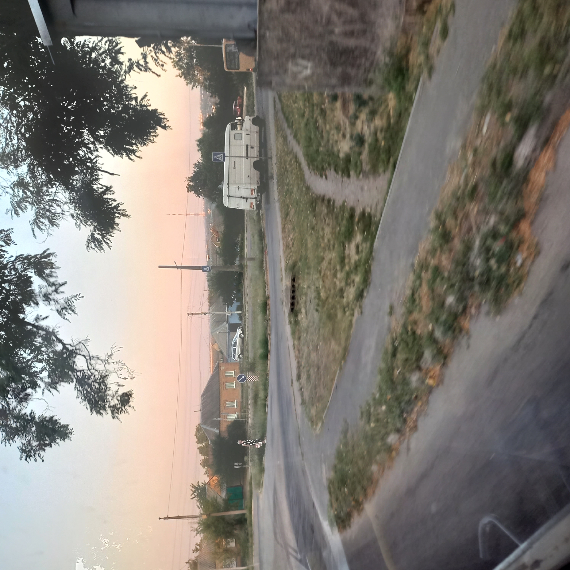 The image shows a street scene in Kropyvnytskyi, Ukraine, likely during either sunrise or sunset due to the warm, diffused light. The foreground is dominated by a paved road and grassy verges. In the middle ground, a woman in a polka dot dress is standing on a sidewalk near a traffic intersection. She is carrying a bag. Behind her, there are houses with tiled roofs and trees. A white van is parked or driving on the road, and a yellow bus is visible in the distance. A pedestrian crossing sign is present. The background reveals more houses and trees, with a hazy horizon. The sky is a gradient of light pink and blue.