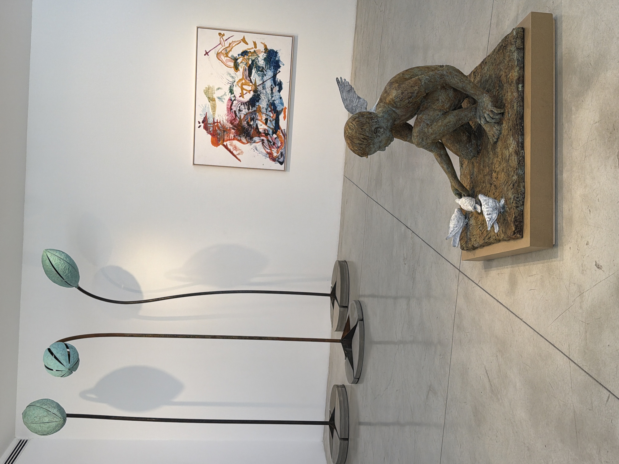An indoor exhibition space, likely a gallery in Tel Aviv, Israel, features a collection of artworks. In the foreground, a bronze sculpture of a winged boy kneels on a textured base, extending his hand towards two small birds, with another bird nearby. The sculpture rests on a simple wooden plinth. To the left, three tall, abstract sculptures with green, pod-like tops stand on circular concrete bases, casting shadows on the white wall behind them. In the background, a colorful, abstract painting with bold brushstrokes in shades of orange, blue, and red hangs on the same white wall. The floor is a polished concrete surface.
