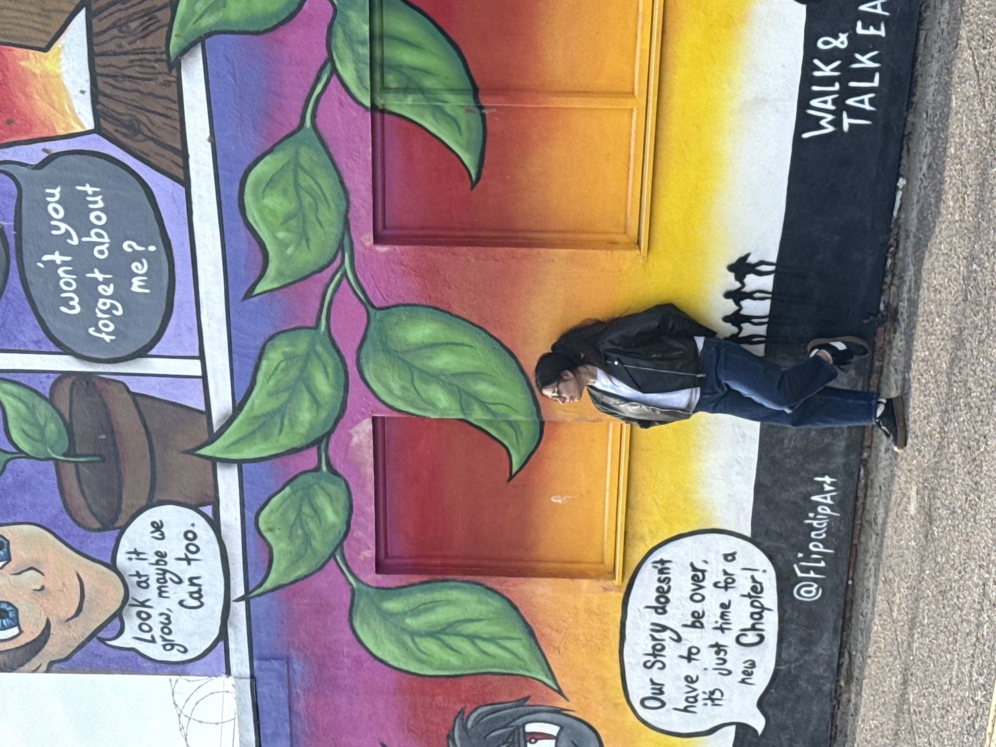 The image shows a woman standing in front of a vibrant mural. She is wearing a black jacket, a white shirt, and blue jeans, and is looking towards the left. The mural consists of comic-strip style panels with colorful backgrounds of purple, red, orange, and yellow. One panel features a plant in a pot with the speech bubble, "Look at it grow, maybe we can too," and another panel with the speech bubble, "won't you forget about me?". A plant with green leaves runs vertically along the wall. Another panel displays a speech bubble with the words "Our Story doesn't have to be over, it's just time for a new Chapter!". The mural also includes the text "@FlipadipArt" and "WALK & TALK EA" on a black background with white text, and silhouettes of people walking. The location is Gorleston-on-Sea, United Kingdom.