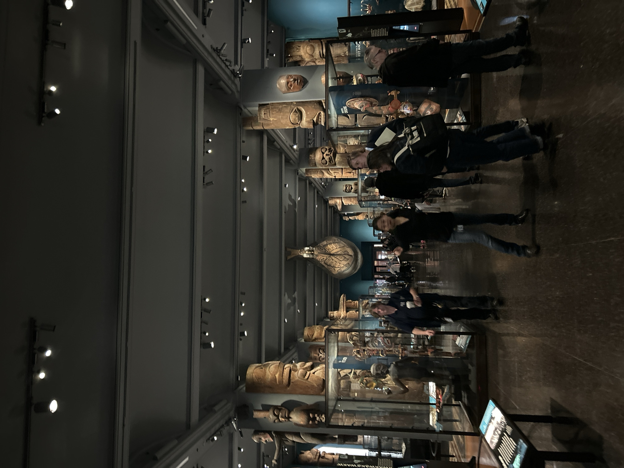 The image shows the interior of the American Museum of Natural History in New York City. The scene focuses on an exhibit of totem poles and other indigenous art displayed in glass cases. Several people are seen observing the exhibits, some wearing backpacks and dark clothing. The museum's architecture features a high ceiling with rows of track lighting. The floor is dark and polished. Display panels provide information about the artifacts.