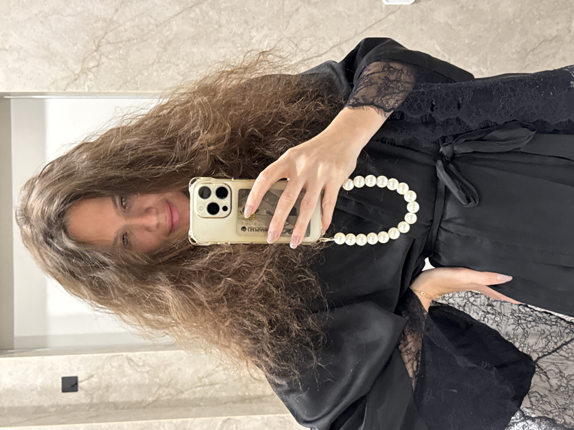 An indoor mirror selfie depicts a woman with long, voluminous, wavy brown hair, looking directly into the camera with a slight smile. She is wearing a black satin-like robe or dress, detailed with delicate black lace trim on the sleeves. Her left hand holds a light gold-colored smartphone with a clear case, from which a decorative white pearl strap dangles around her wrist. Her nails are manicured with a light, possibly glittery or ombre, polish.
Through the clear phone case, a card is visible with Cyrillic text, clearly showing "СБЕРБАНК" (SBERBANK), a prominent Russian financial institution, which aligns with the provided location context of Moscow, Russia. The background, visible in the mirror reflection, consists of light-toned, possibly beige or gray, marble or stone walls with subtle veining. The setting appears to be a well-lit room, possibly a bathroom or dressing area.