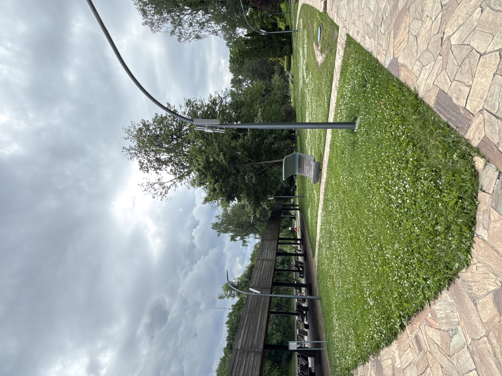 The image depicts an outdoor park scene with a cloudy sky overhead. In the foreground, a stone pathway curves to the right, bordering a lush green lawn dotted with small white flowers. To the left, a long wooden structure with a covered roof, supported by dark posts, stretches into the background. Beneath this structure, rows of benches are visible. A modern-style bench is situated on the lawn to the right of the pathway. Two lampposts, one with a curved arm, are prominent features within the park. Trees form a dense backdrop behind the structures and lawn. The overall impression is one of a tranquil public space. This scene is located in the Ostrov Mechty (Dream Island) Park in Moscow, Russia, specifically in the themed zone "Fairy Tale Forest."