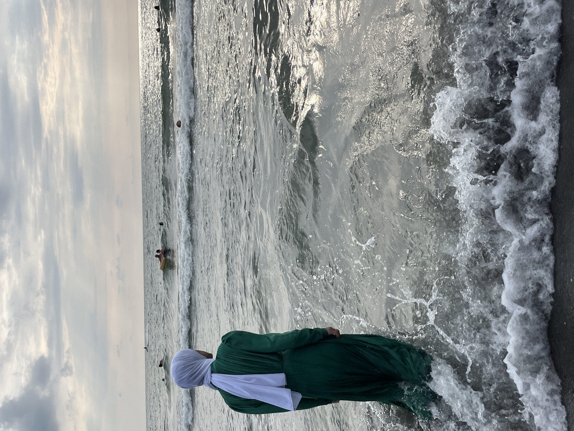 A person wearing a green dress and white hijab stands in the shallow water of the Ureki beach in Georgia, facing the sea. The person's back is to the camera. Waves are breaking around their legs, creating splashes. In the distance, several people are visible in the water, some on inflatables and one on what appears to be a jet ski. The sky is overcast with clouds. The scene appears to be captured during the day.