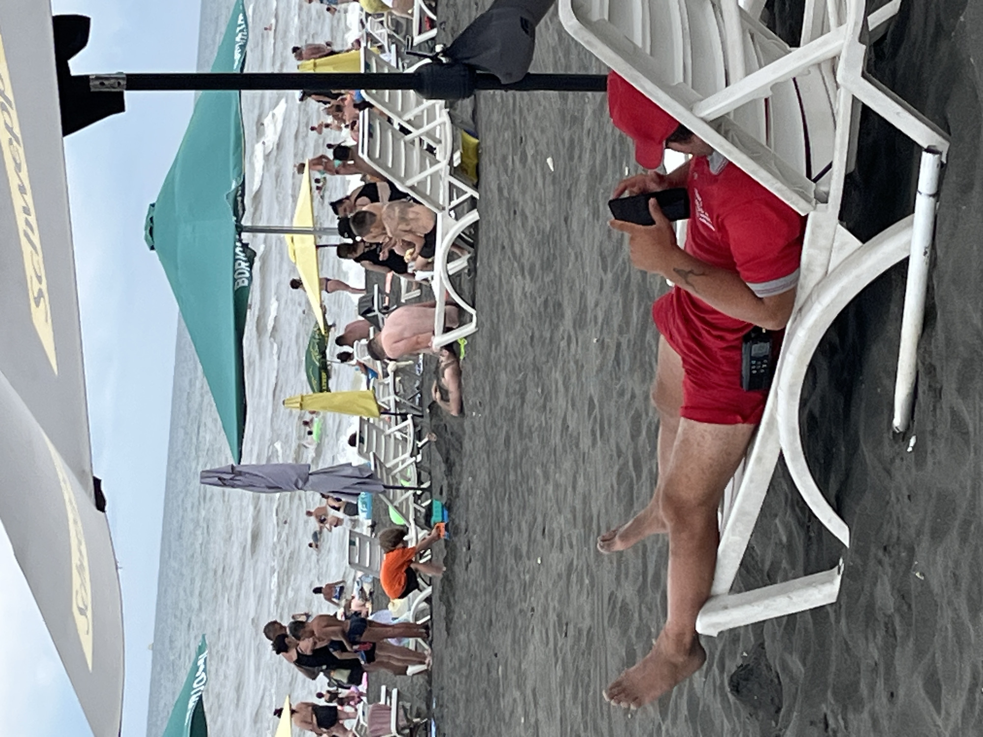 The image depicts a crowded beach scene in Ureki, Georgia. In the foreground, a person lies on a white lounge chair on the dark sand, wearing a red shirt and shorts, and holding a black smartphone. The person's legs are extended, and their feet are visible. In the middle ground, several other individuals are seen relaxing on lounge chairs, with some in the water. Beach umbrellas, including a large white one with a logo, a green one with text, and a yellow one, are set up on the sand. People are scattered across the beach and in the ocean, which appears to have moderate waves. The overall atmosphere suggests a typical summer day at a popular seaside resort.