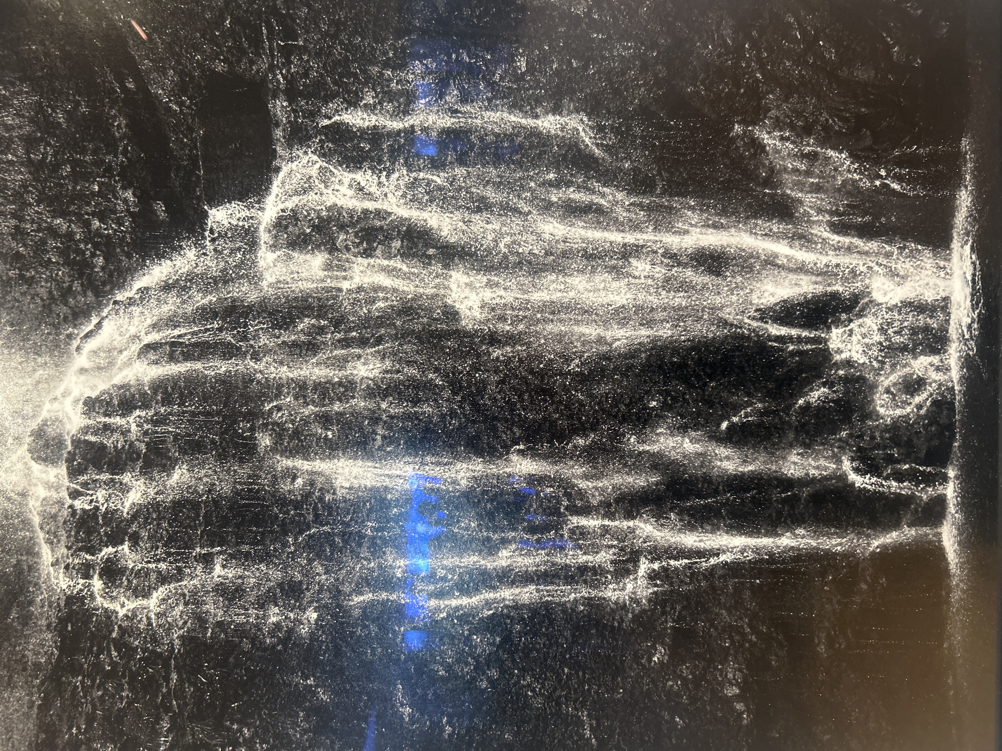 The image shows a black and white photograph of what appears to be a water feature cascading down a rocky surface. The water is rendered as bright white streaks and splashes against a dark background. The overall texture is rough and uneven, suggesting a natural, perhaps rocky, environment. There are no people or other discernible subjects visible. The lighting appears natural, potentially daytime. No specific location in West Hollywood, United States of America can be ascertained from this image.