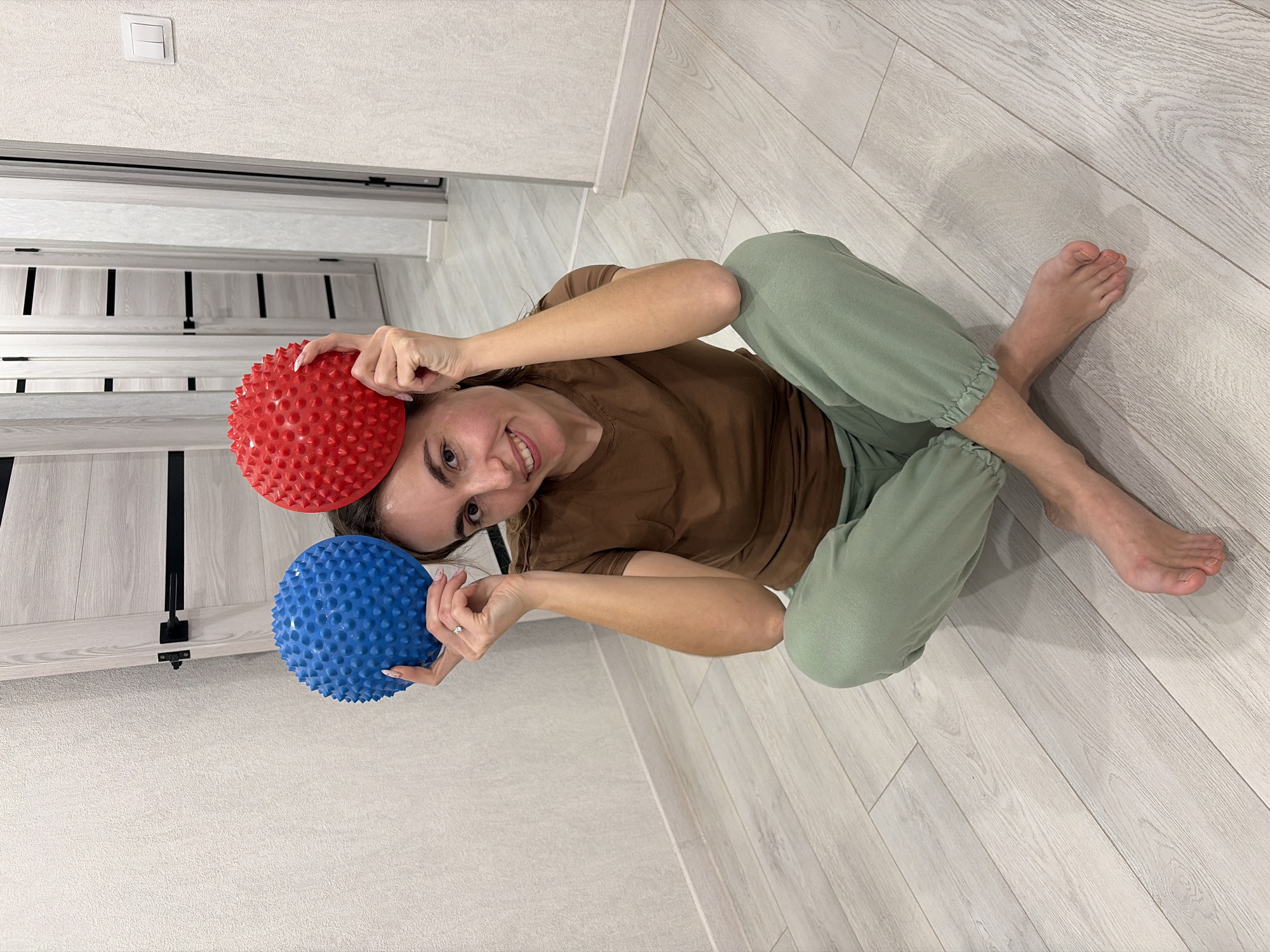 The image shows a woman indoors, sitting cross-legged on a light-colored wooden floor. She is wearing a brown t-shirt and light green sweatpants. She is holding a red spiky ball above her head with her right hand and a blue spiky ball next to her head with her left hand. The woman is smiling. A light switch is visible on the wall in the upper left corner. A door with a black latch is also visible. The image was taken in Sarapul, Russia.